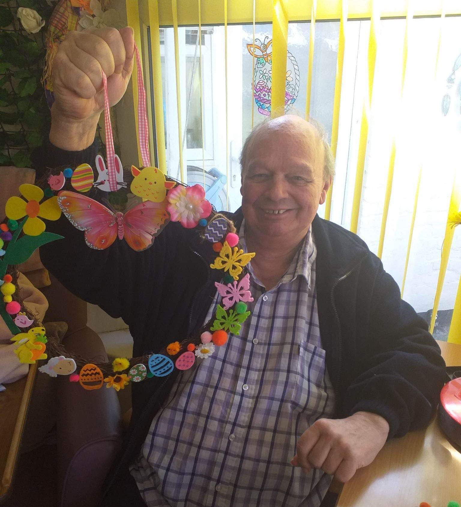 A smiling resident at Wisteria Stoke holds up a colourful Easter-themed wreath made with pom-poms, flowers, and foam shapes during a craft activity.