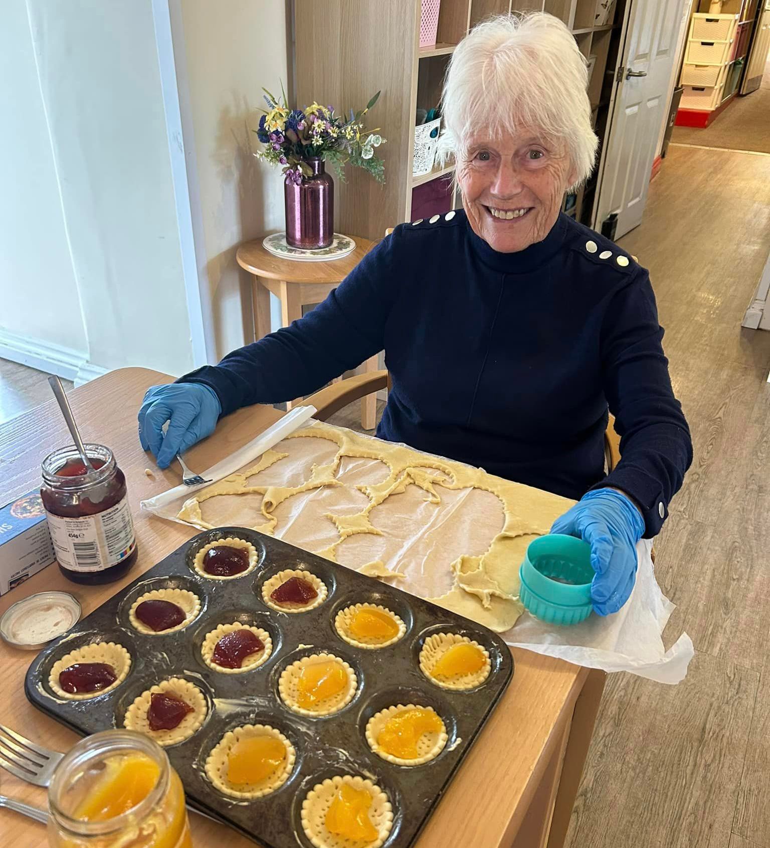 A smiling resident at Wisteria Plymstock prepares jam tarts at a table, surrounded by pastry, jars of jam, and a tray of filled tart cases.