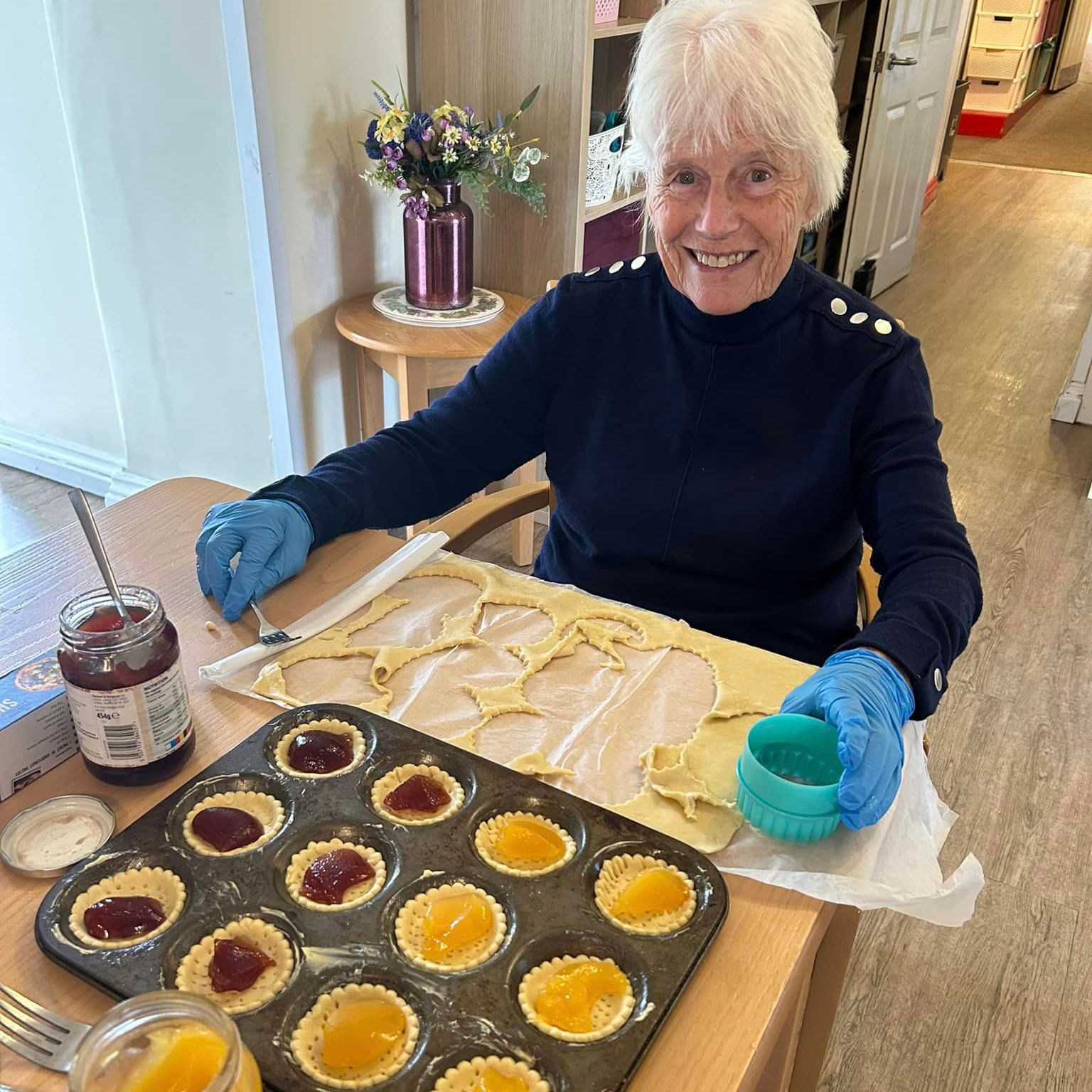 A smiling resident at Wisteria Plymstock prepares jam tarts at a table, surrounded by pastry, jars of jam, and a tray of filled tart cases.