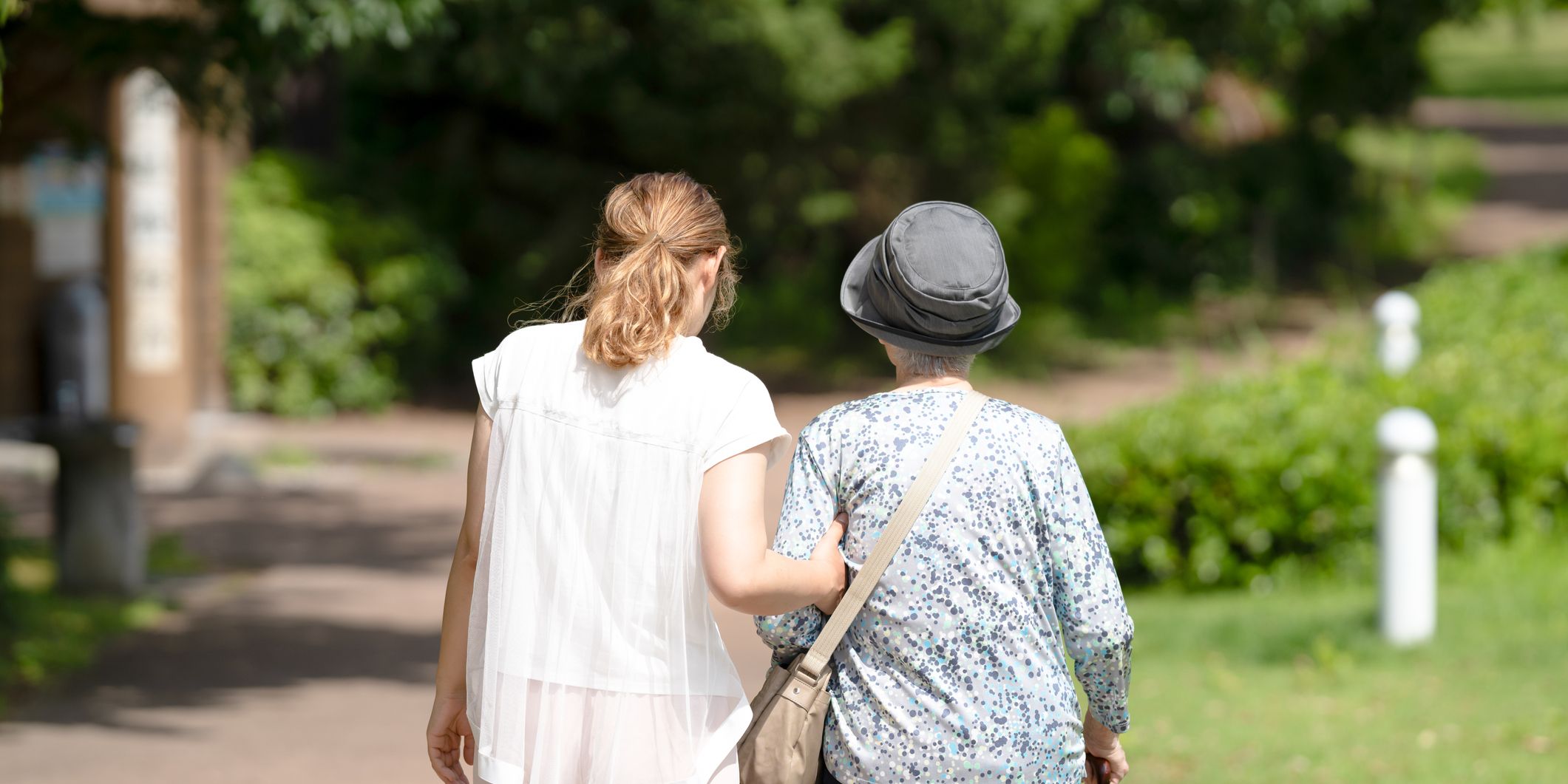 A younger woman gently supports an older woman with a walking stick as they walk together along a sunlit path in a green park.