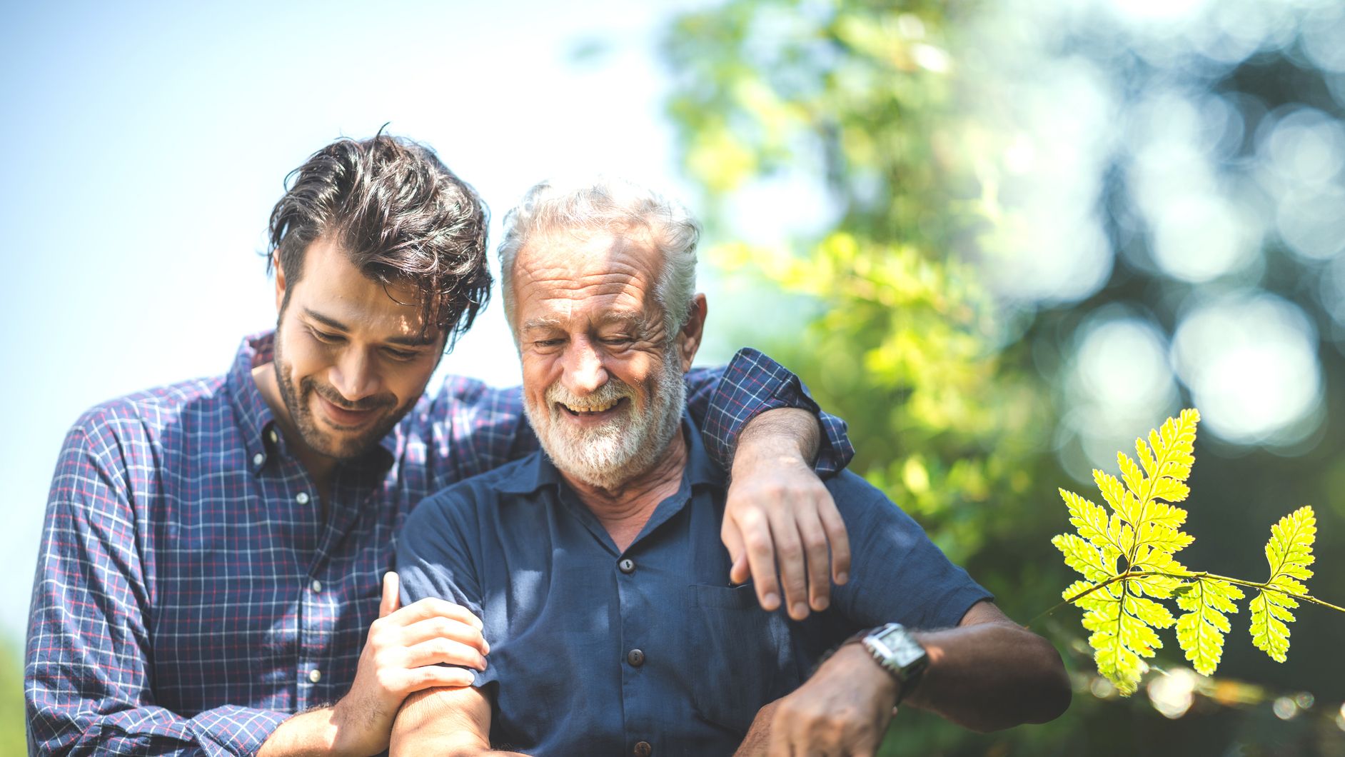 Younger and older man smiling together outdoors, with one arm around the other in soft sunlight and blurred greenery behind.