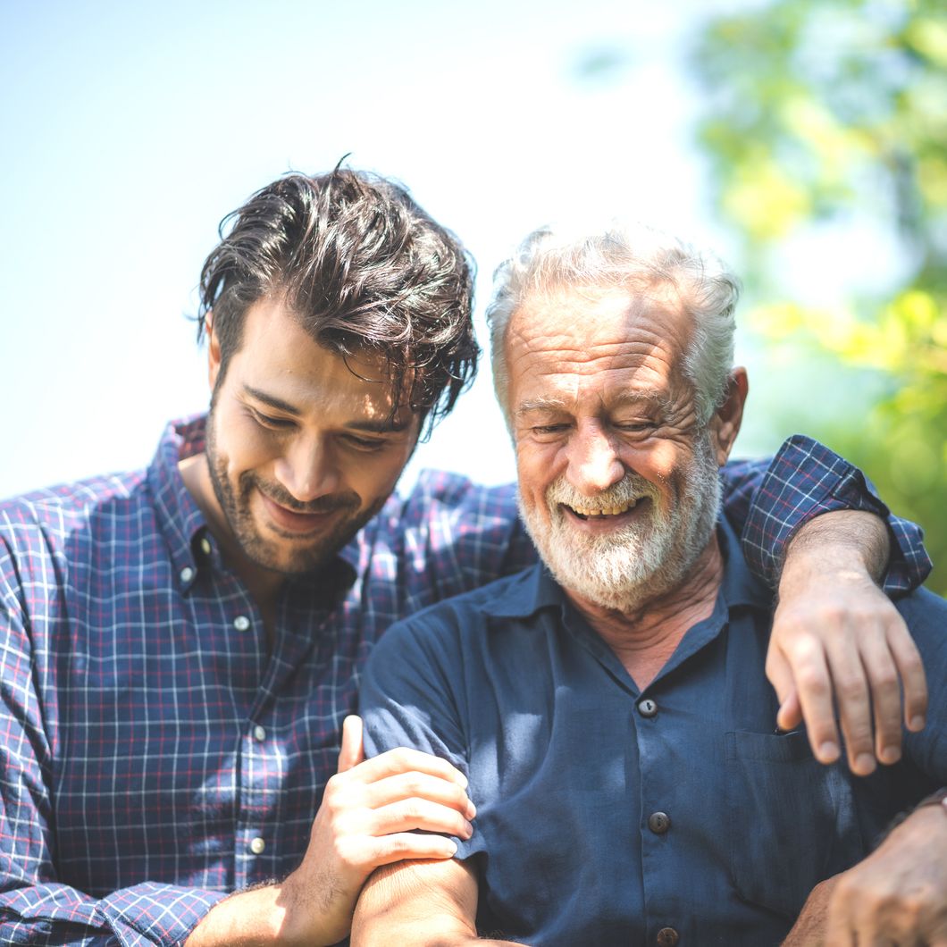 Younger and older man smiling together outdoors, with one arm around the other in soft sunlight and blurred greenery behind.