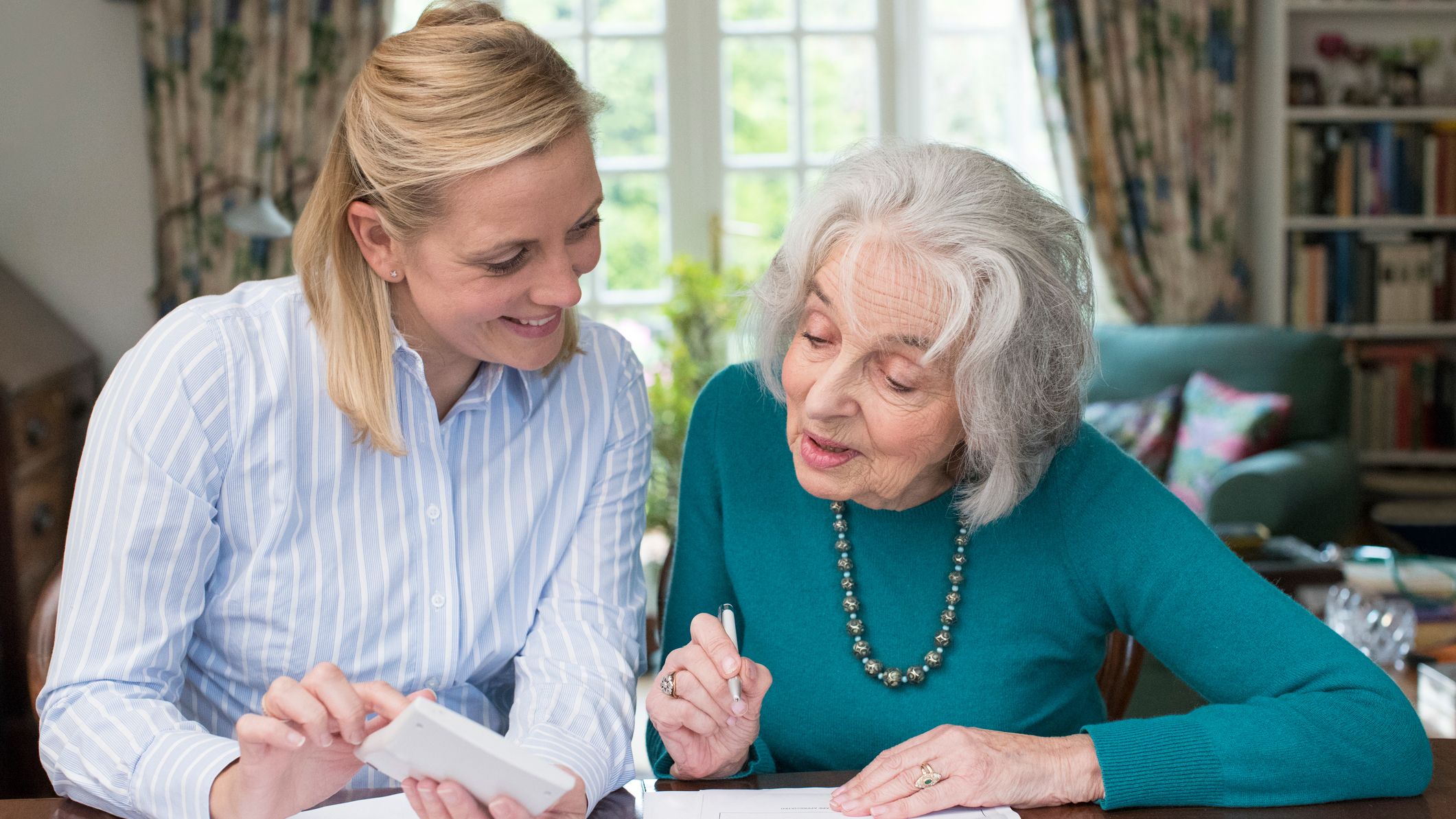 A younger woman and an older woman review paperwork at a table, while one holds a calculator-like device in a bright living room.