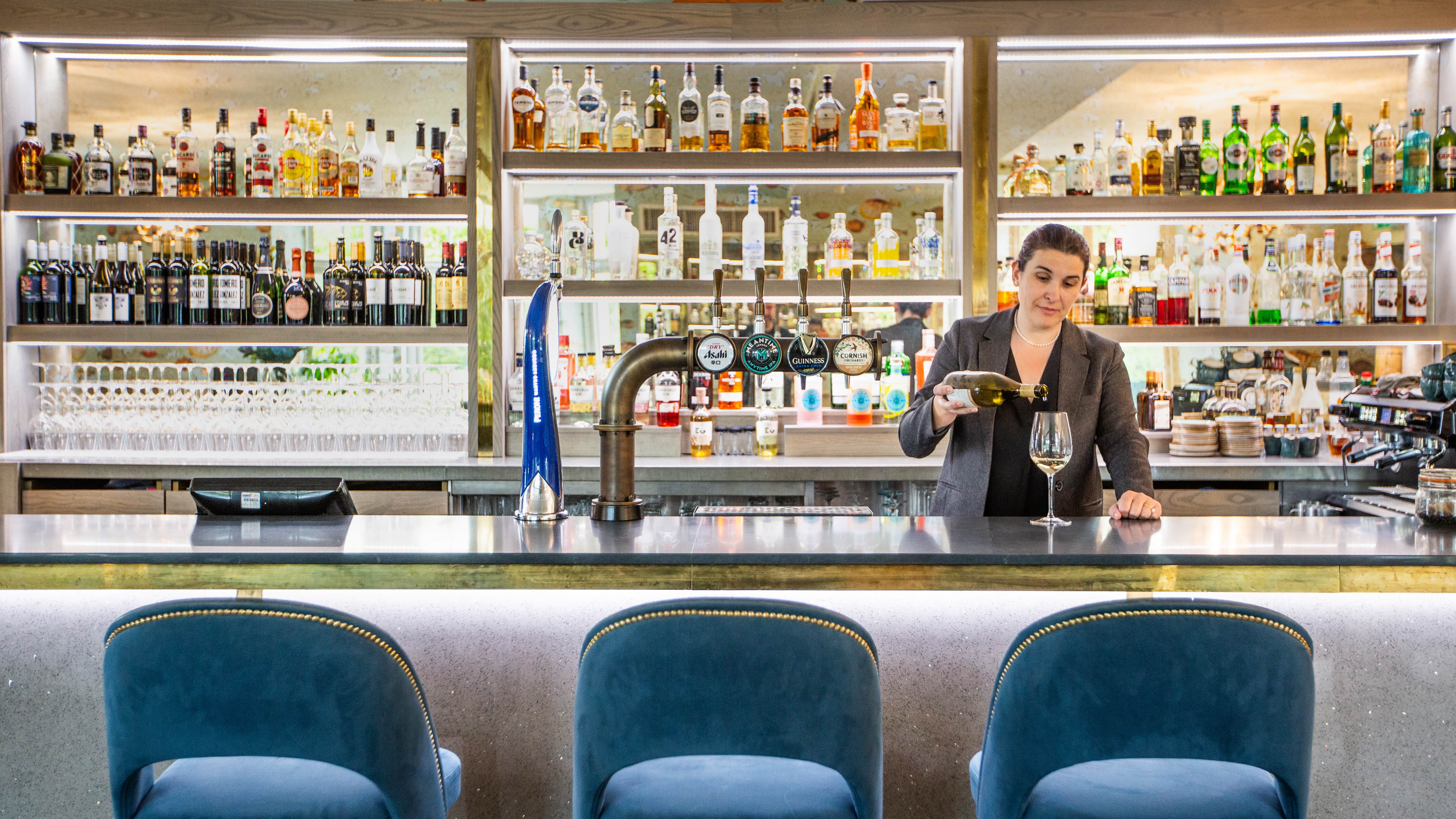 Lady pouring glass of wine behind bar, with three blue chairs tucked in front of the bar