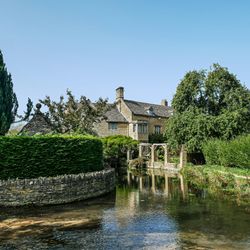 Traditional Cotswold stone cottage beside a calm stream and lush gardens under a clear blue sky