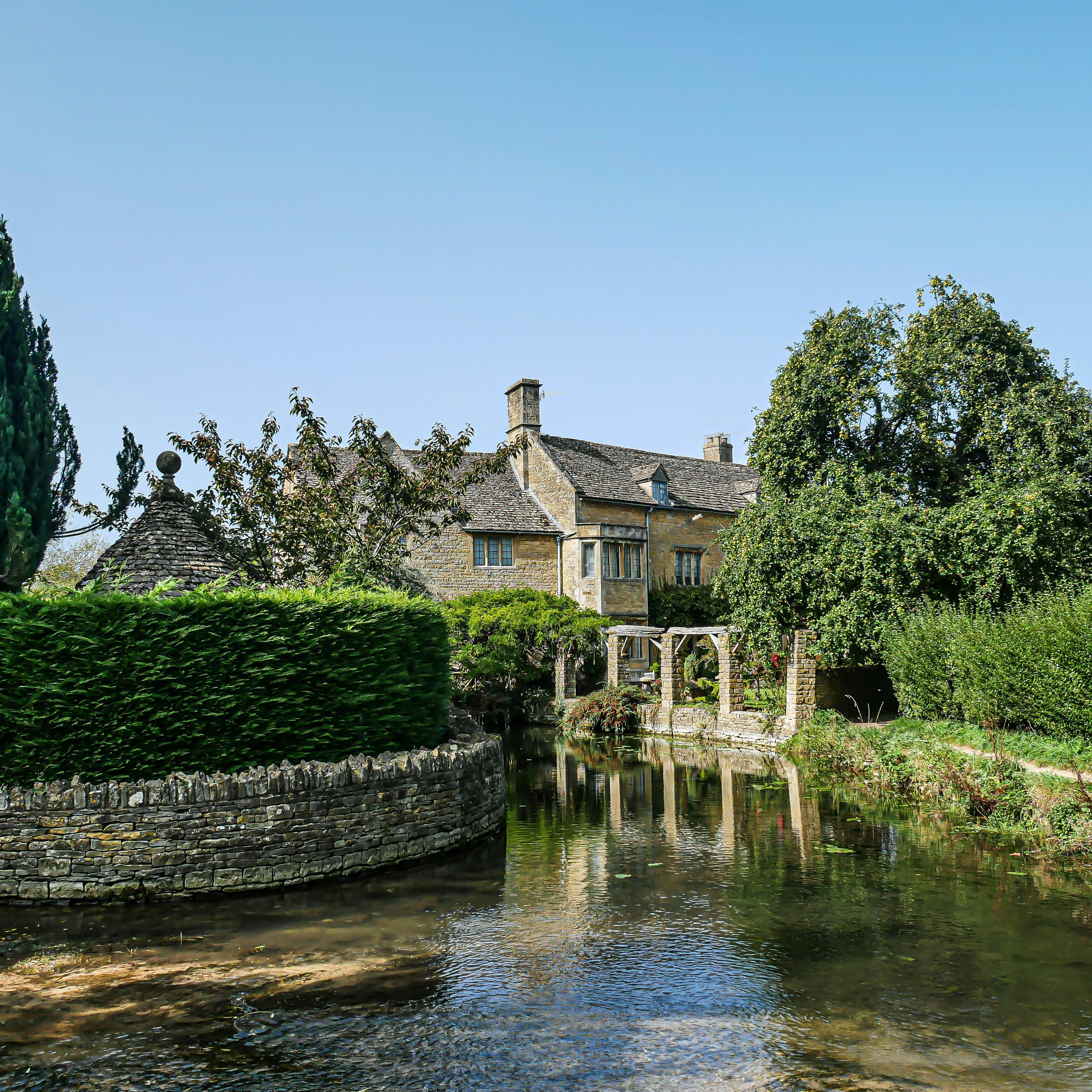 Traditional Cotswold stone cottage beside a calm stream and lush gardens under a clear blue sky