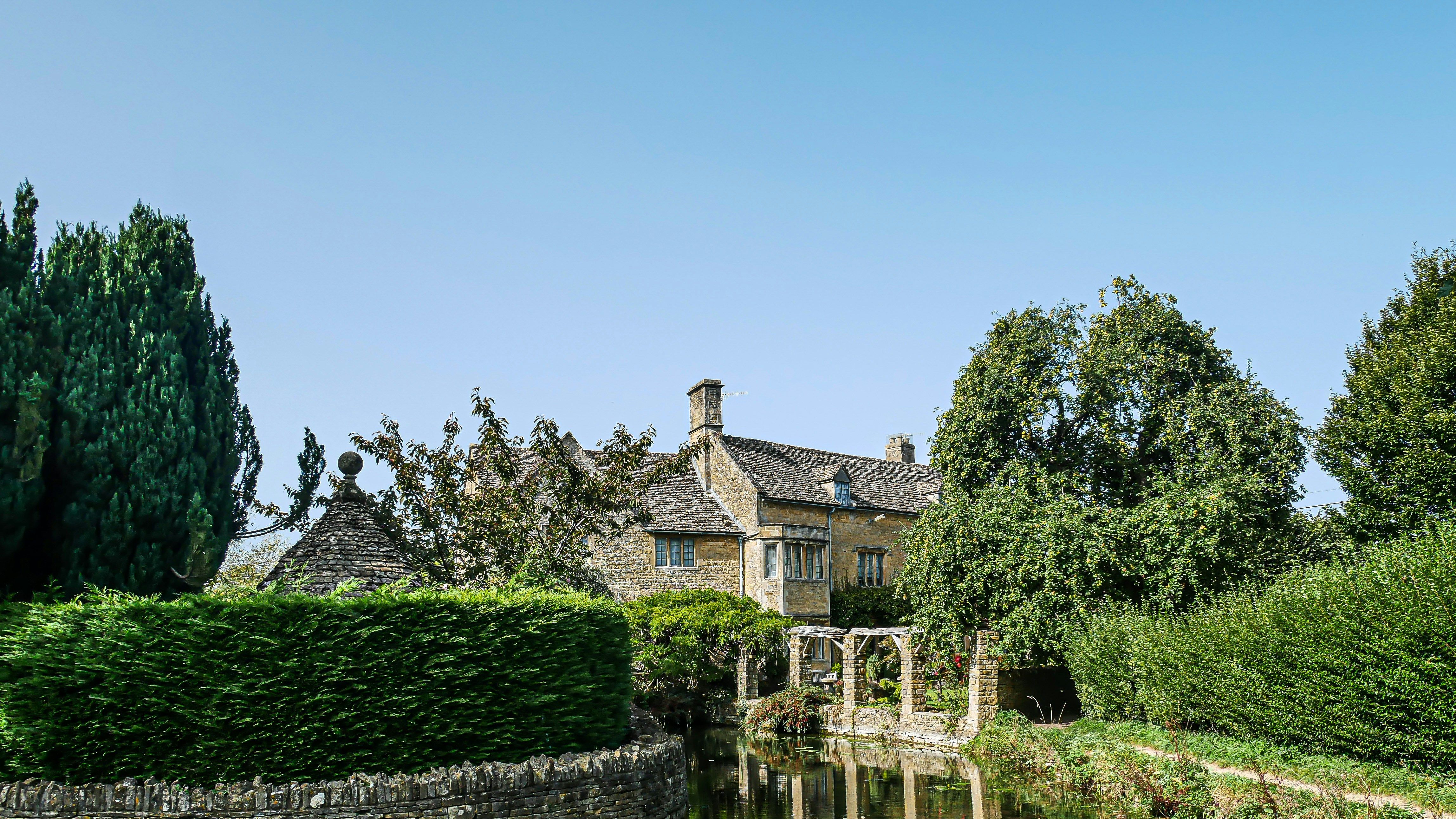 Traditional Cotswold stone cottage beside a calm stream and lush gardens under a clear blue sky