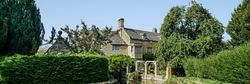 Traditional Cotswold stone cottage beside a calm stream and lush gardens under a clear blue sky