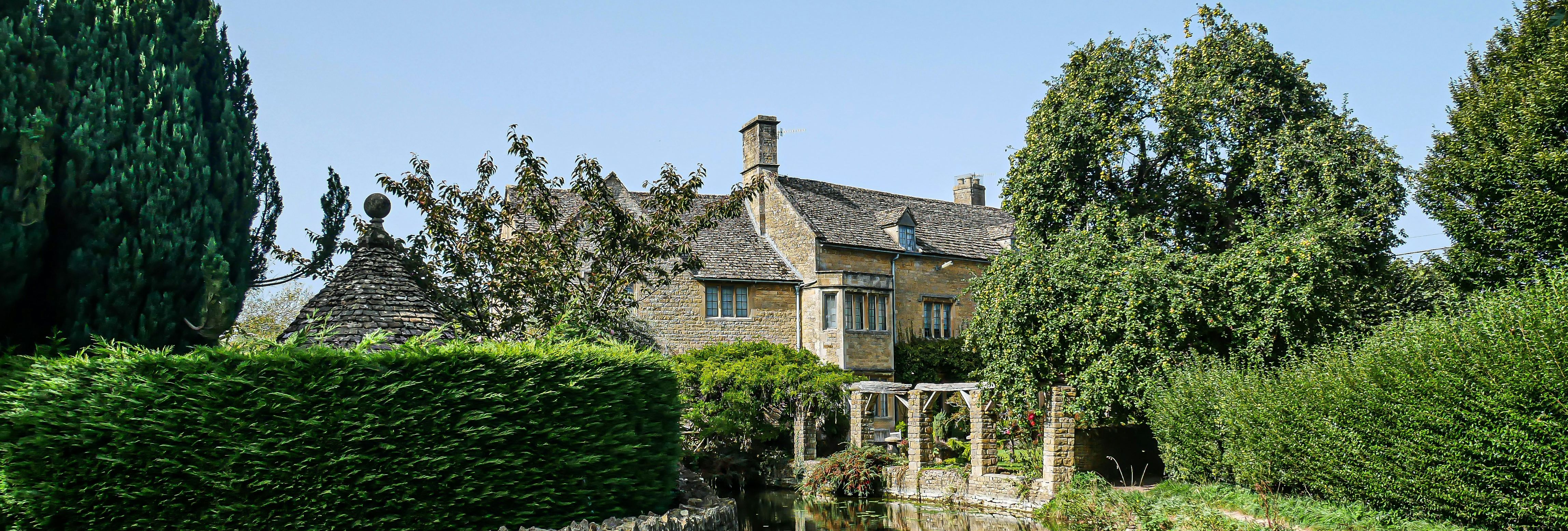Traditional Cotswold stone cottage beside a calm stream and lush gardens under a clear blue sky