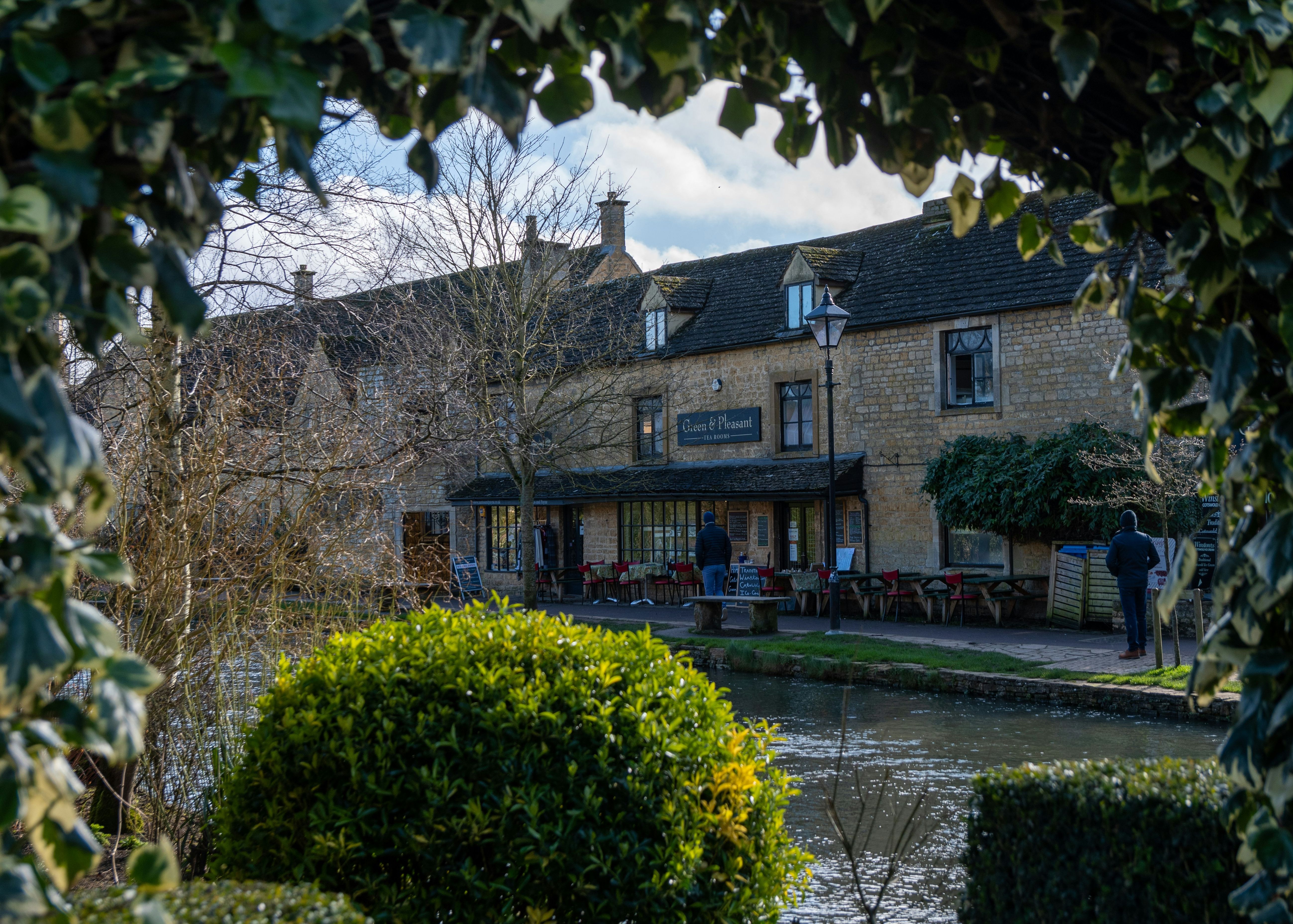 Cotswold village shopfront and canal in Bourton-on-the-Water, framed by greenery