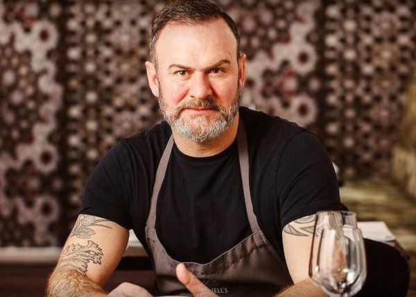 A bearded man wearing a black shirt and apron, seated at a table with a wine glass in front of him.