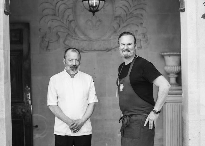Two men in chef attire standing under an ornate archway