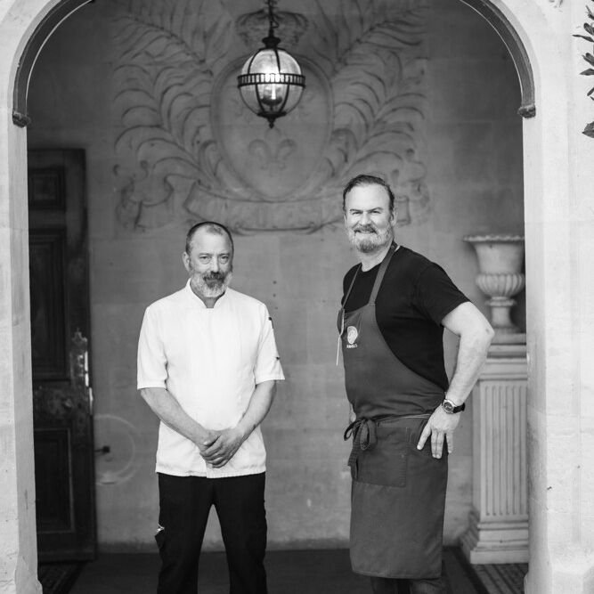 Two men in chef attire standing under an ornate archway
