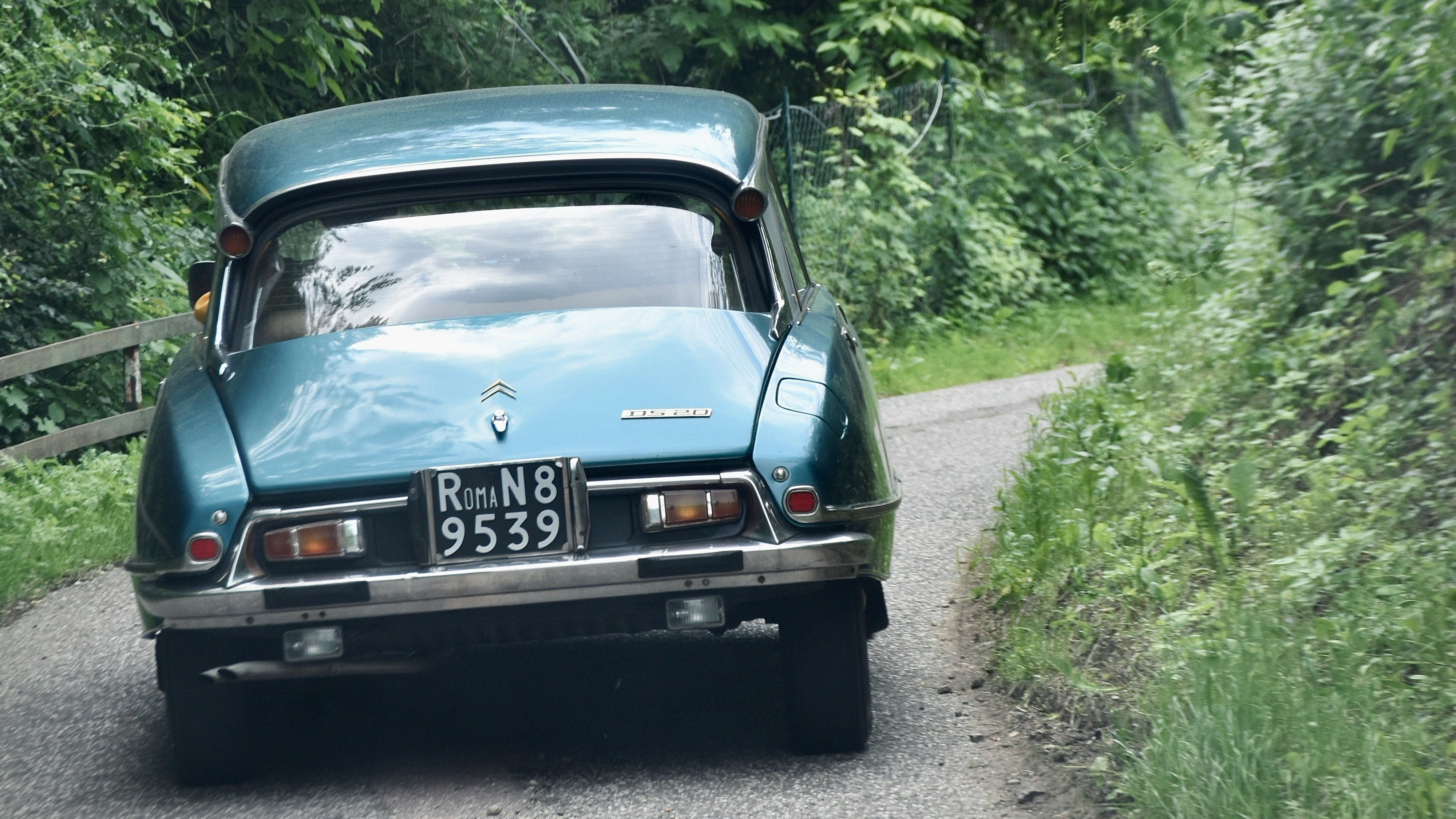 Vintage blue Citroën car driving on a narrow, winding road surrounded by lush green trees and foliage.