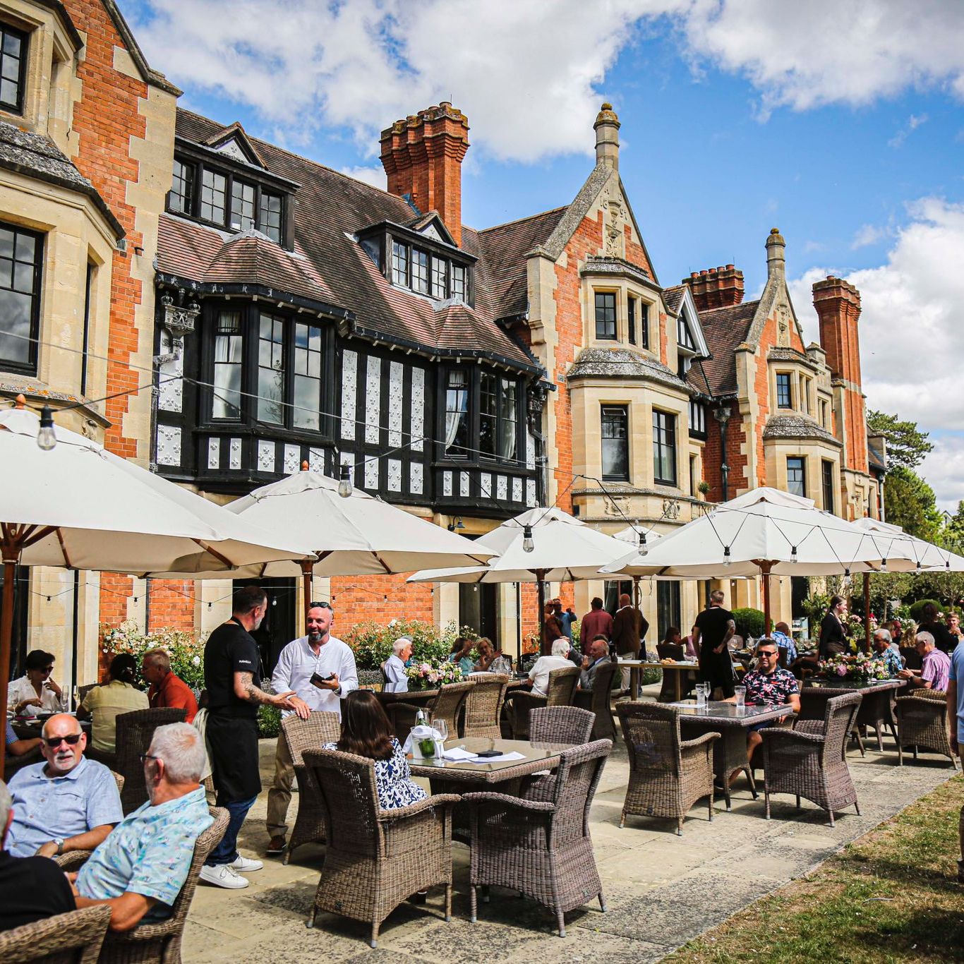 People dining outdoors under white umbrellas in front of a large historic building with chimneys and gabled roofs.