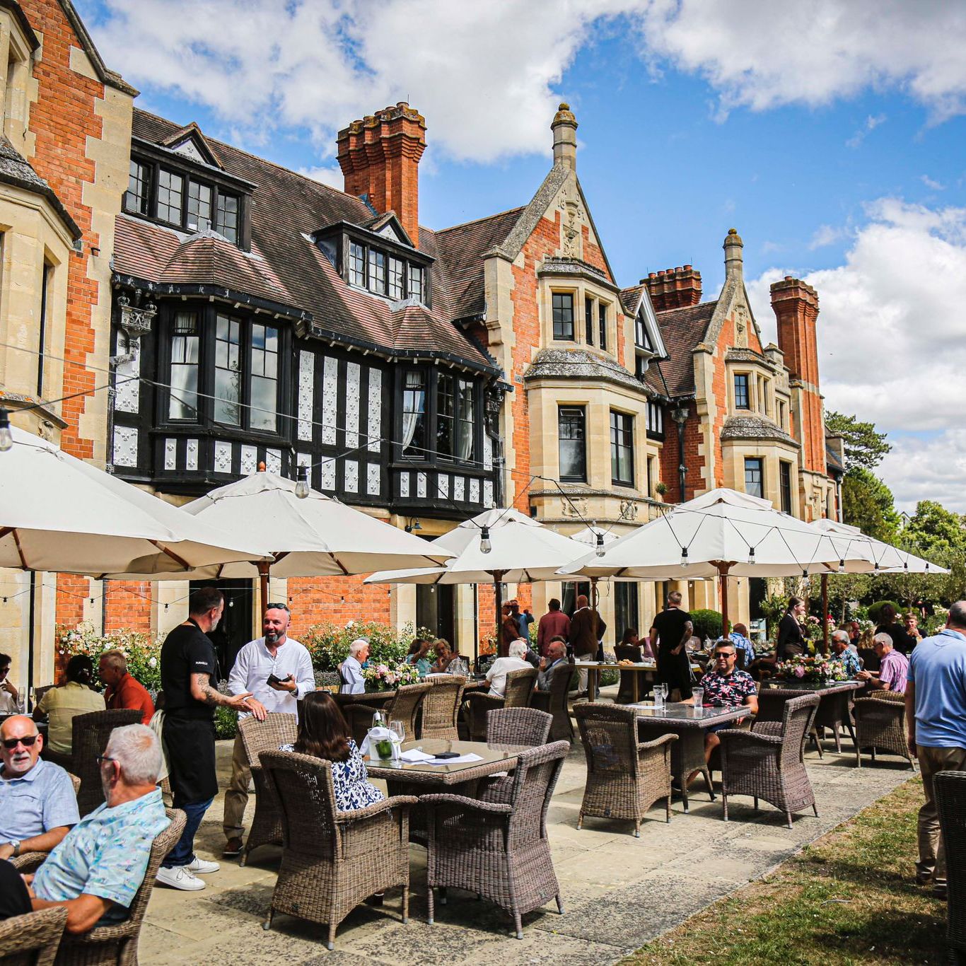 People dining outdoors under white umbrellas in front of a large historic building with chimneys and gabled roofs.