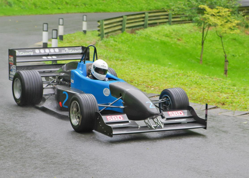 Blue and black single-seater race car on a wet road, driven by a person wearing a white helmet