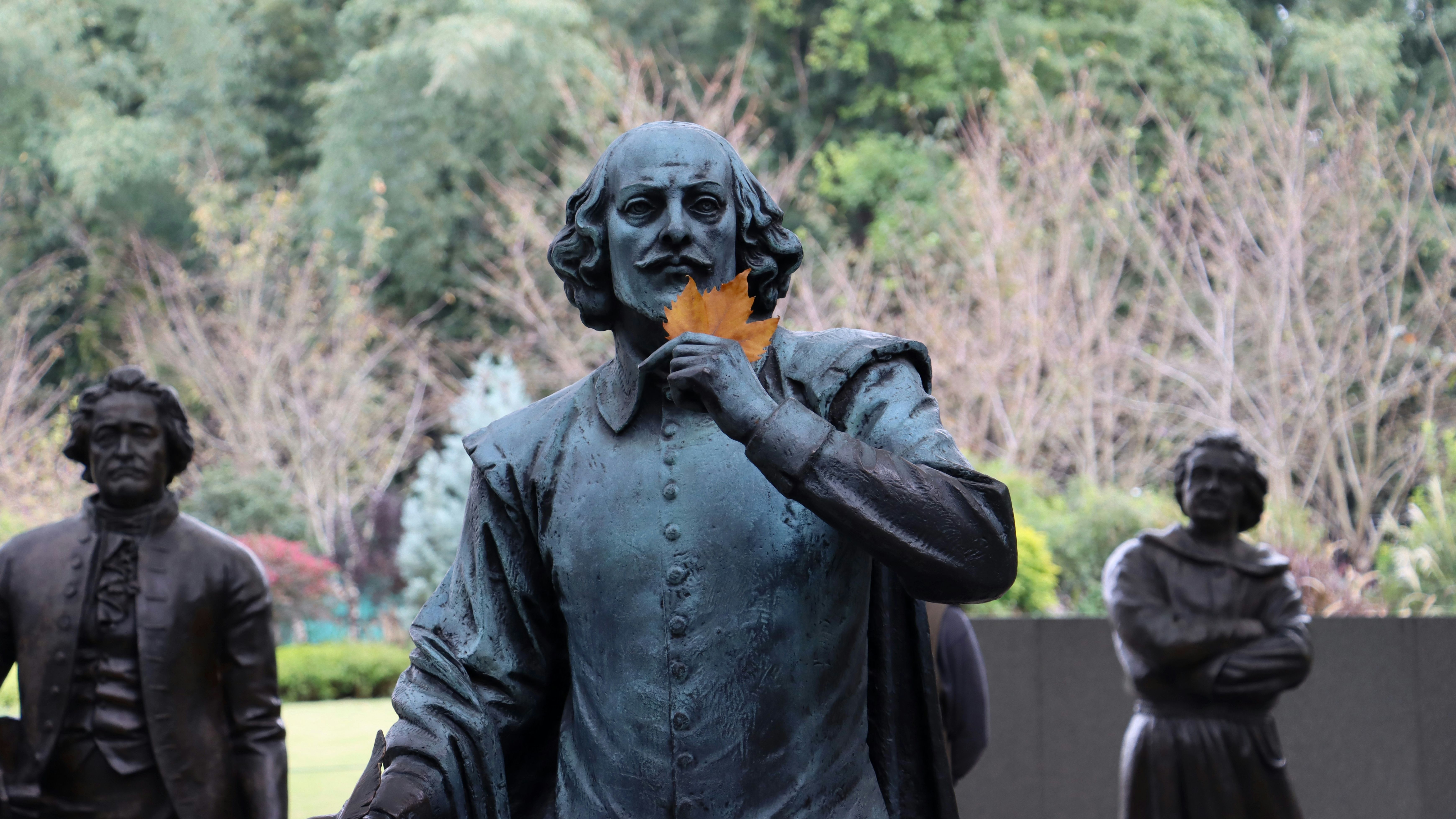 Statue of William Shakespeare holding a leaf to his face, with other statues in the background and trees behind them
