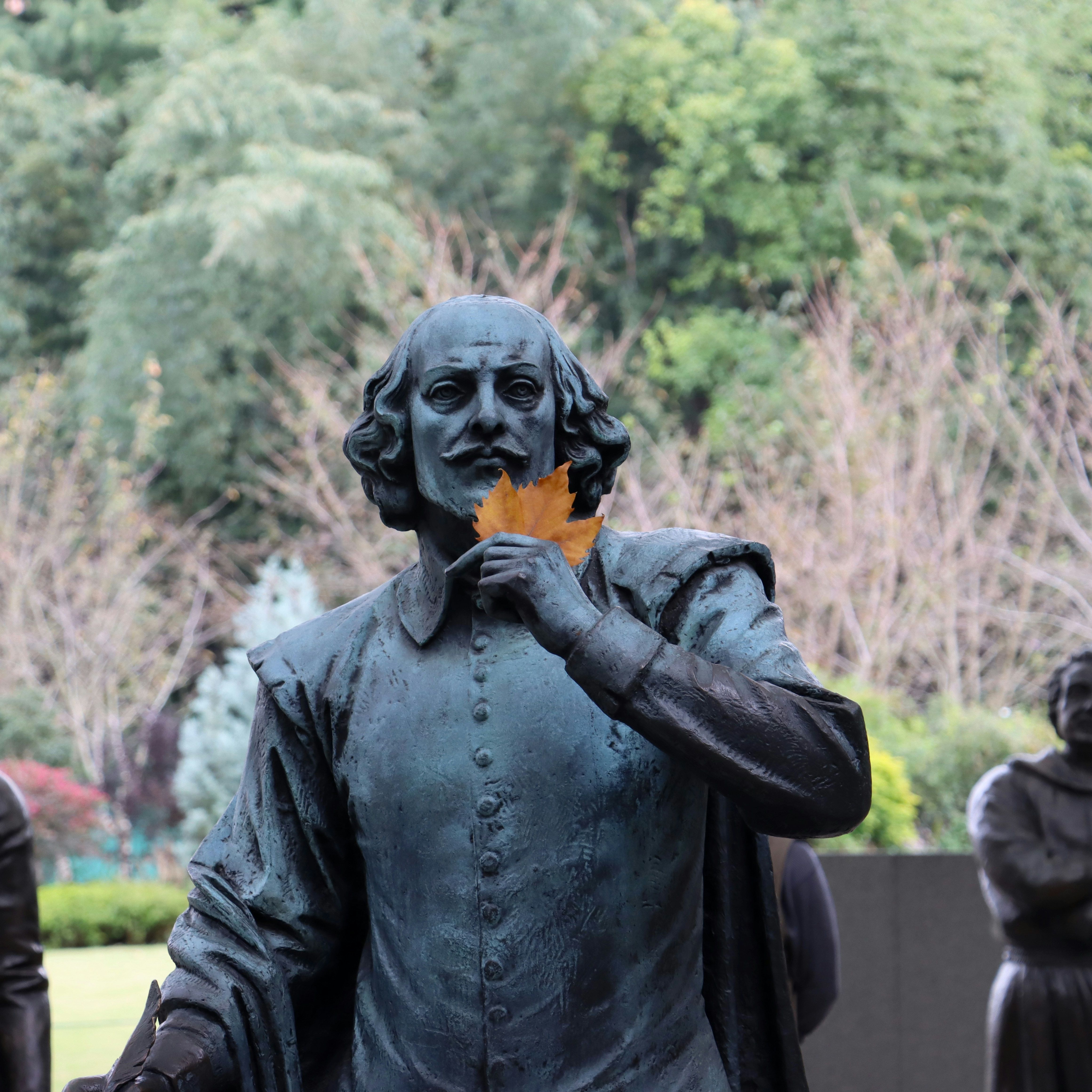 Statue of William Shakespeare holding a leaf to his face, with other statues in the background and trees behind them