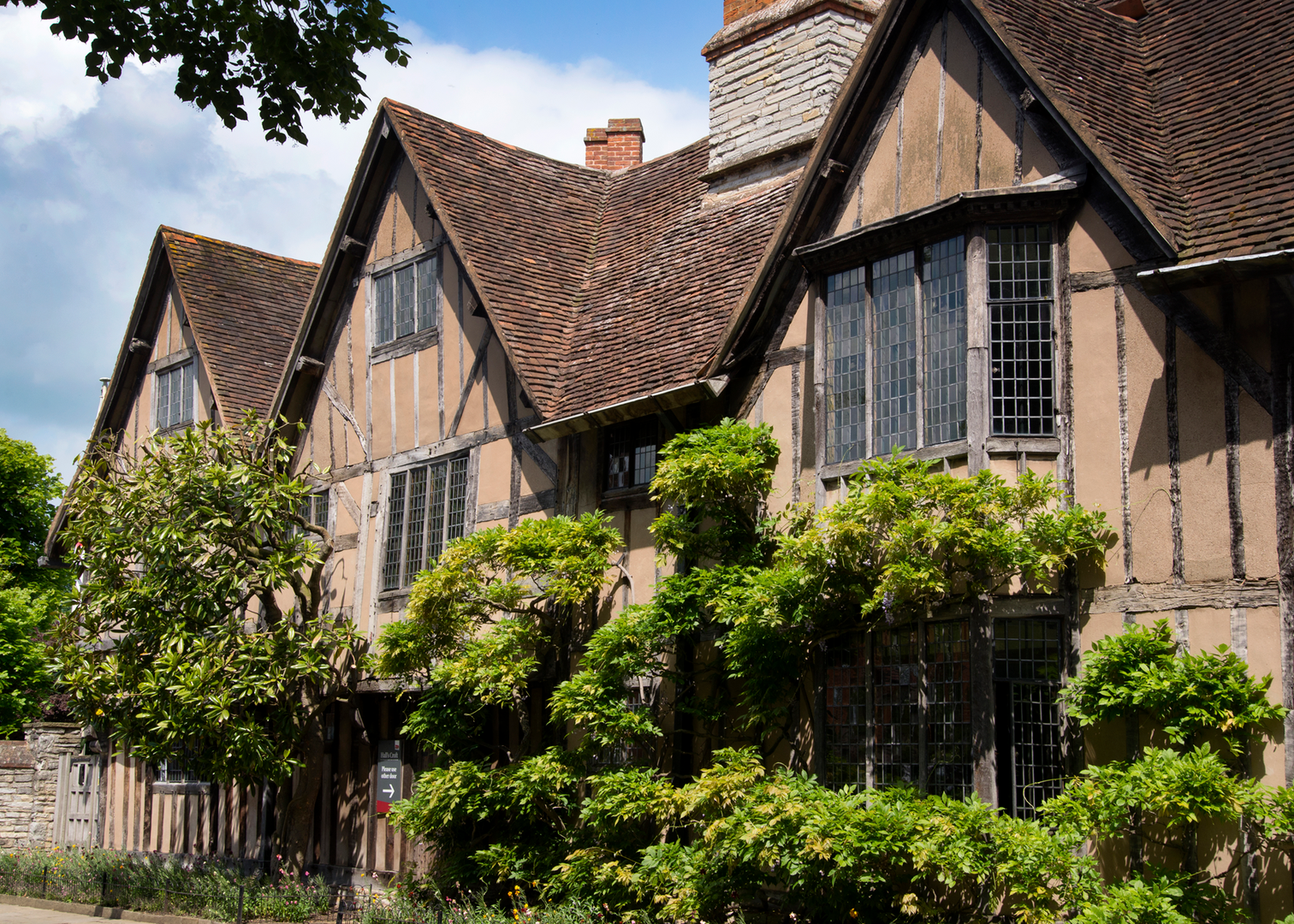Historic half-timbered building with leaded glass windows and a sloped tiled roof, surrounded by greenery.