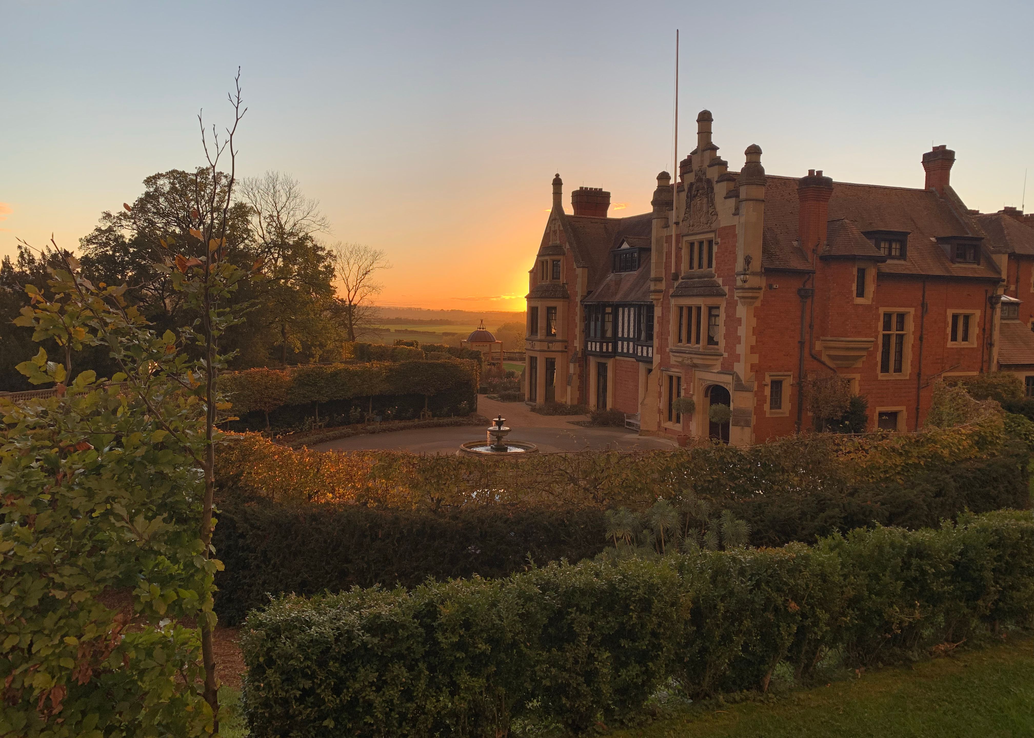 Historic mansion with brick exterior and decorative trim at sunset, surrounded by gardens and a fountain.