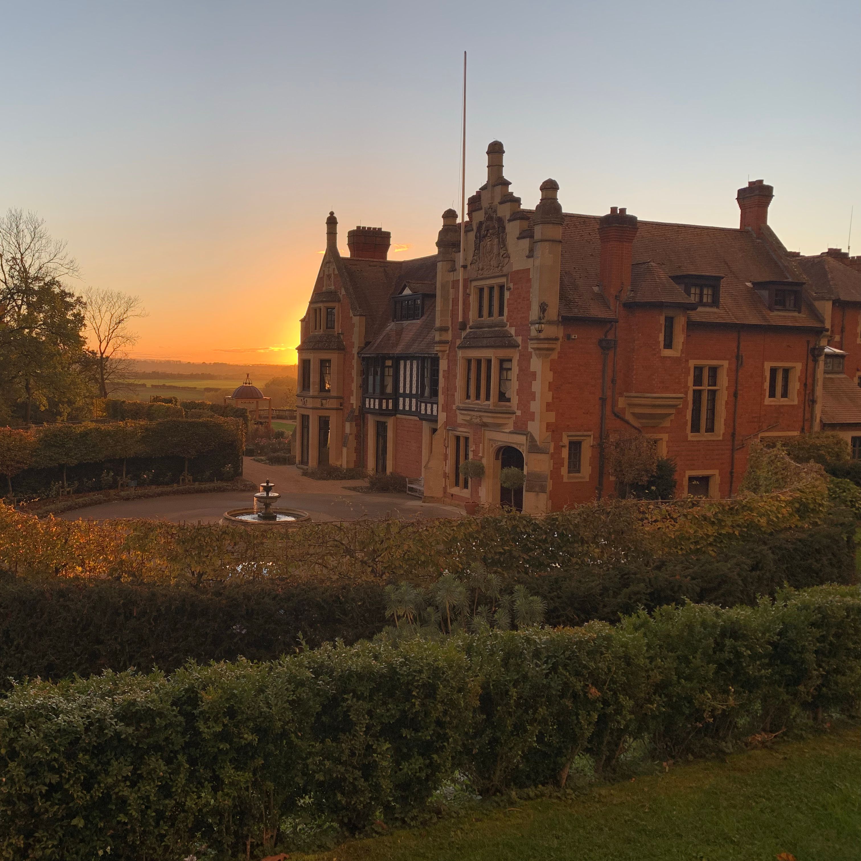 Historic mansion with brick exterior and decorative trim at sunset, surrounded by gardens and a fountain.