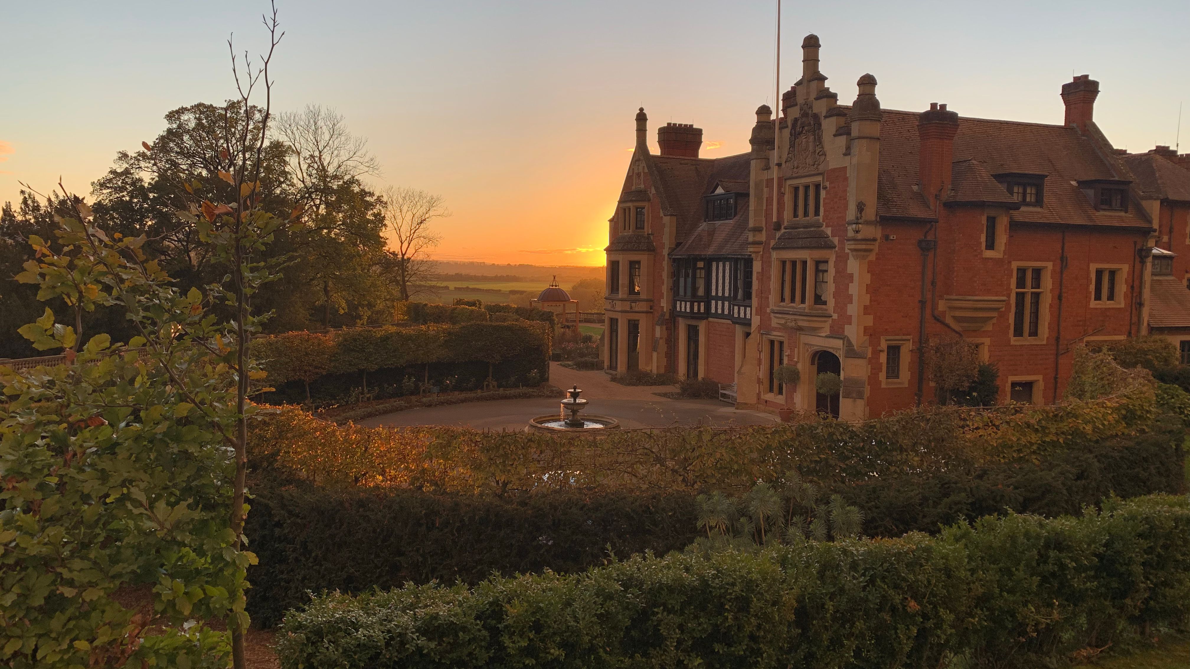 Historic mansion with brick exterior and decorative trim at sunset, surrounded by gardens and a fountain.