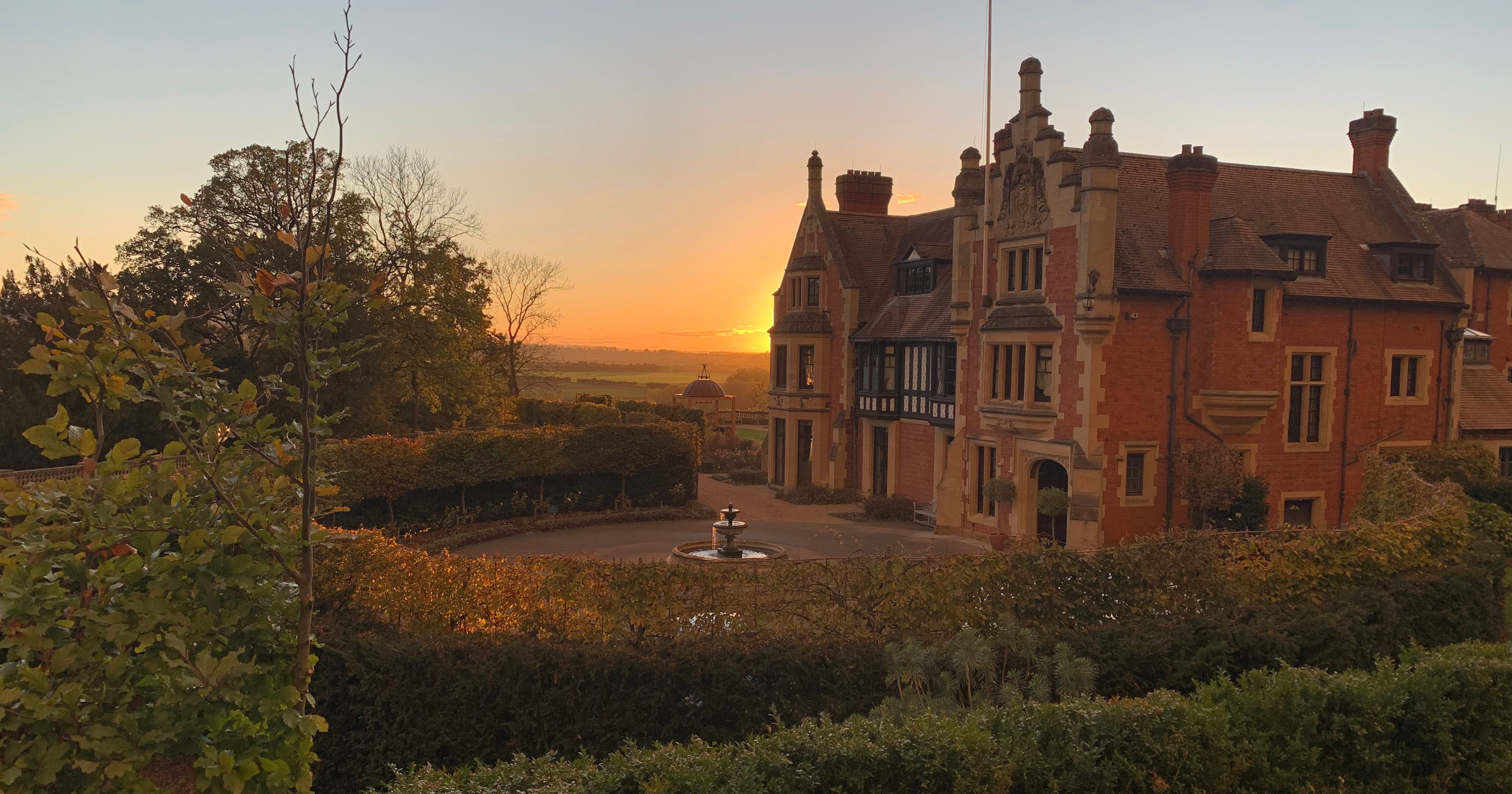Historic mansion with brick exterior and decorative trim at sunset, surrounded by gardens and a fountain.