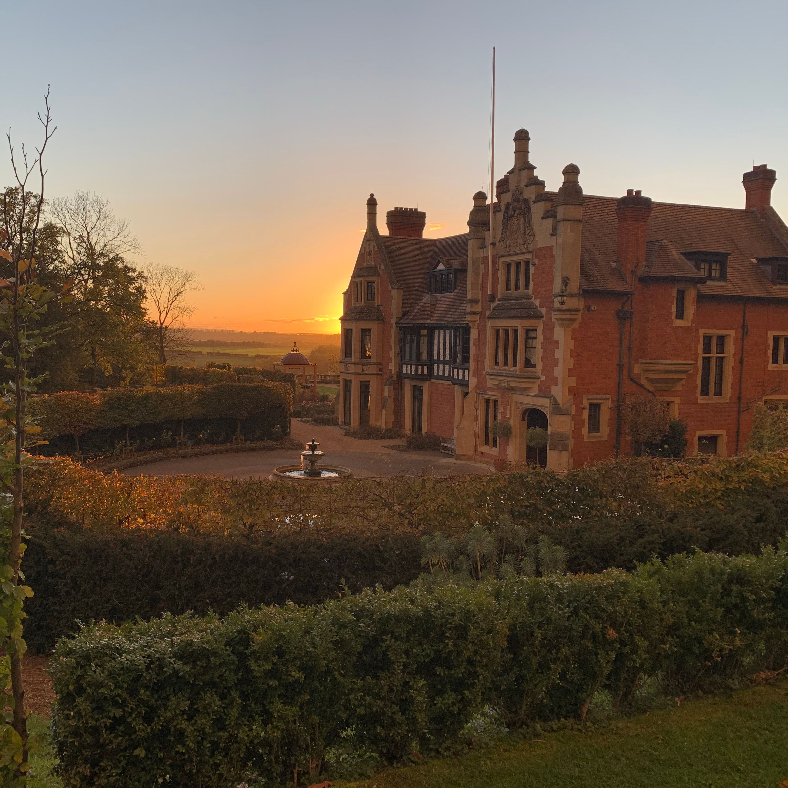 Historic mansion with brick exterior and decorative trim at sunset, surrounded by gardens and a fountain.