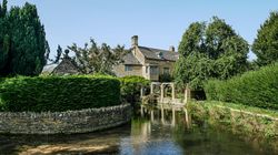 Stone house near a river surrounded by lush greenery and trees under a clear blue sky