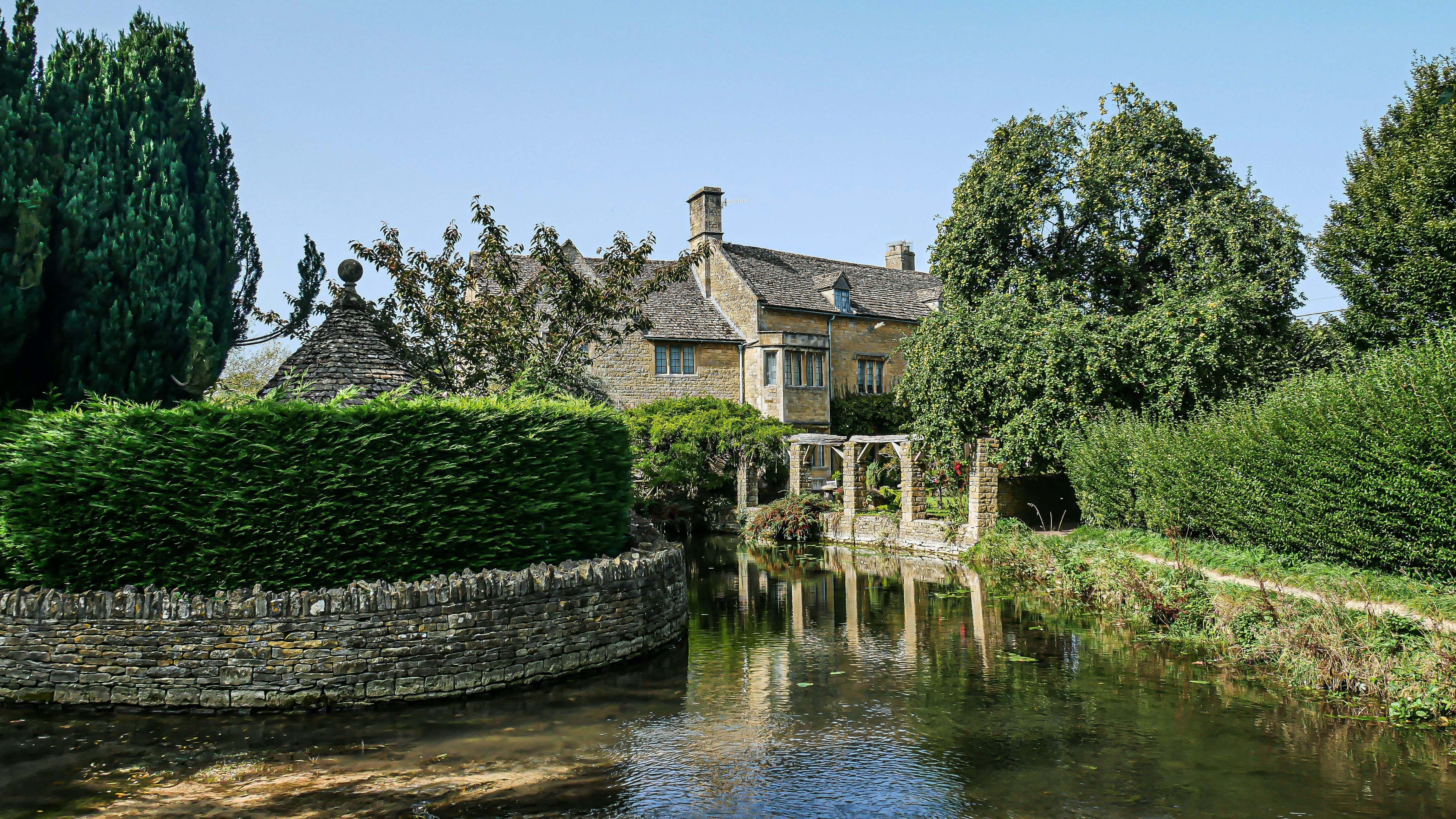 Stone house near a river surrounded by lush greenery and trees under a clear blue sky