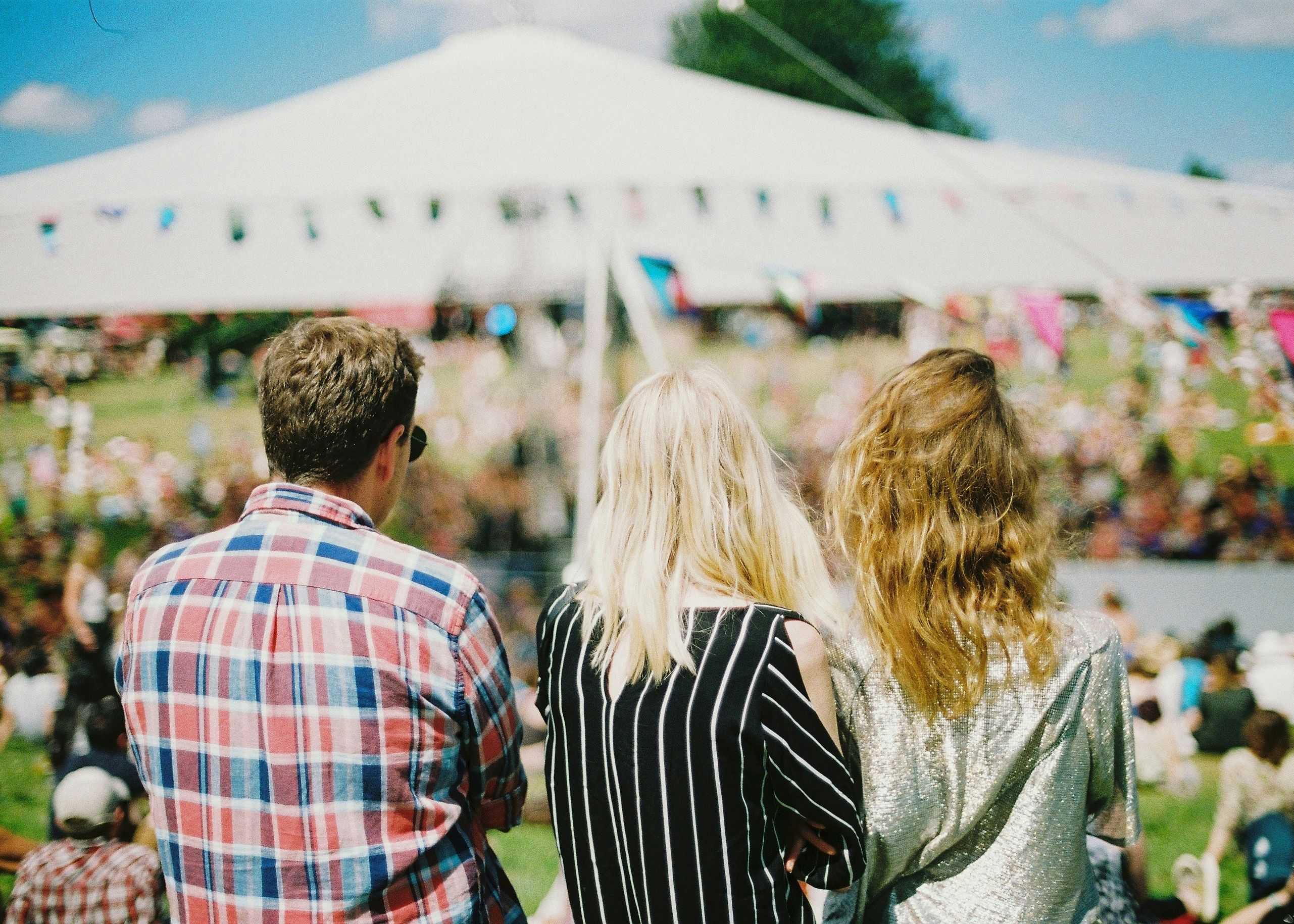 Three people from behind overlooking a lively outdoor festival with a large tent in the background.