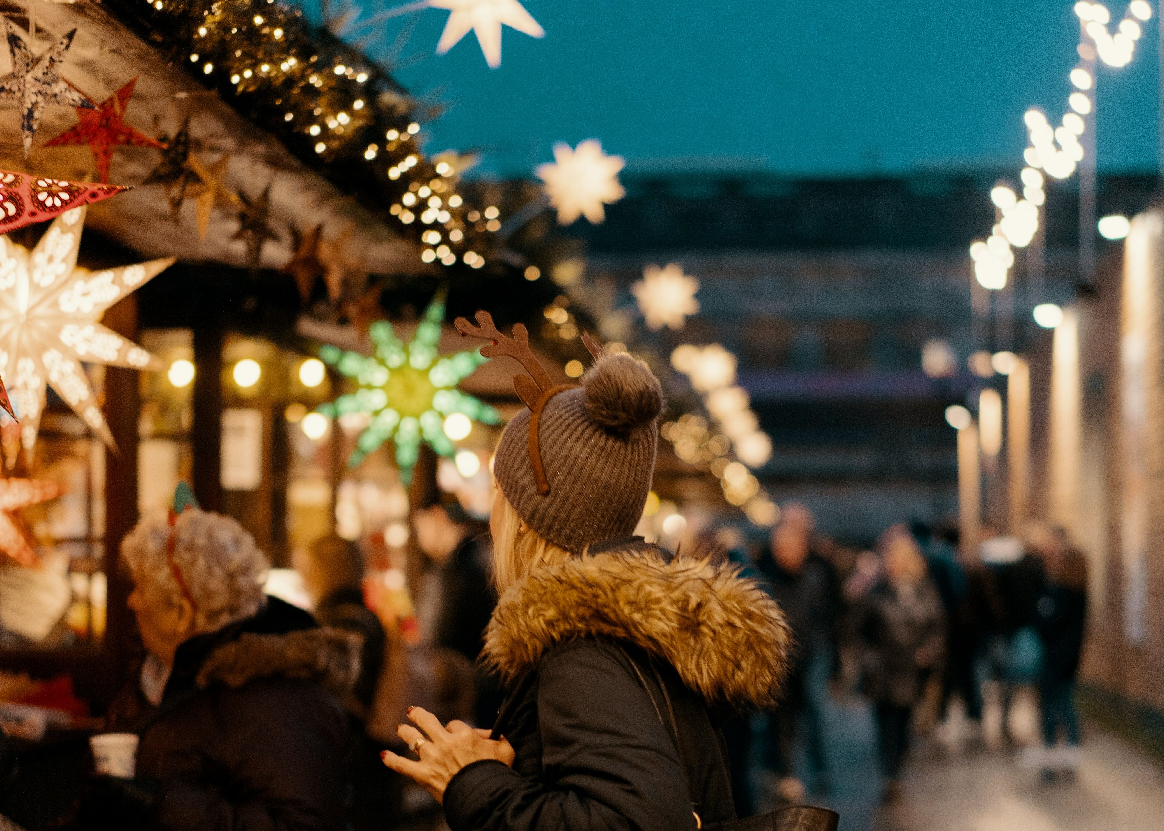 Person wearing a coat and reindeer antlers headband holding a cup at an outdoor market decorated with star-shaped lights