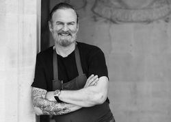 Smiling man with a beard and tattoos wearing an apron, standing with arms crossed by a stone archway.
