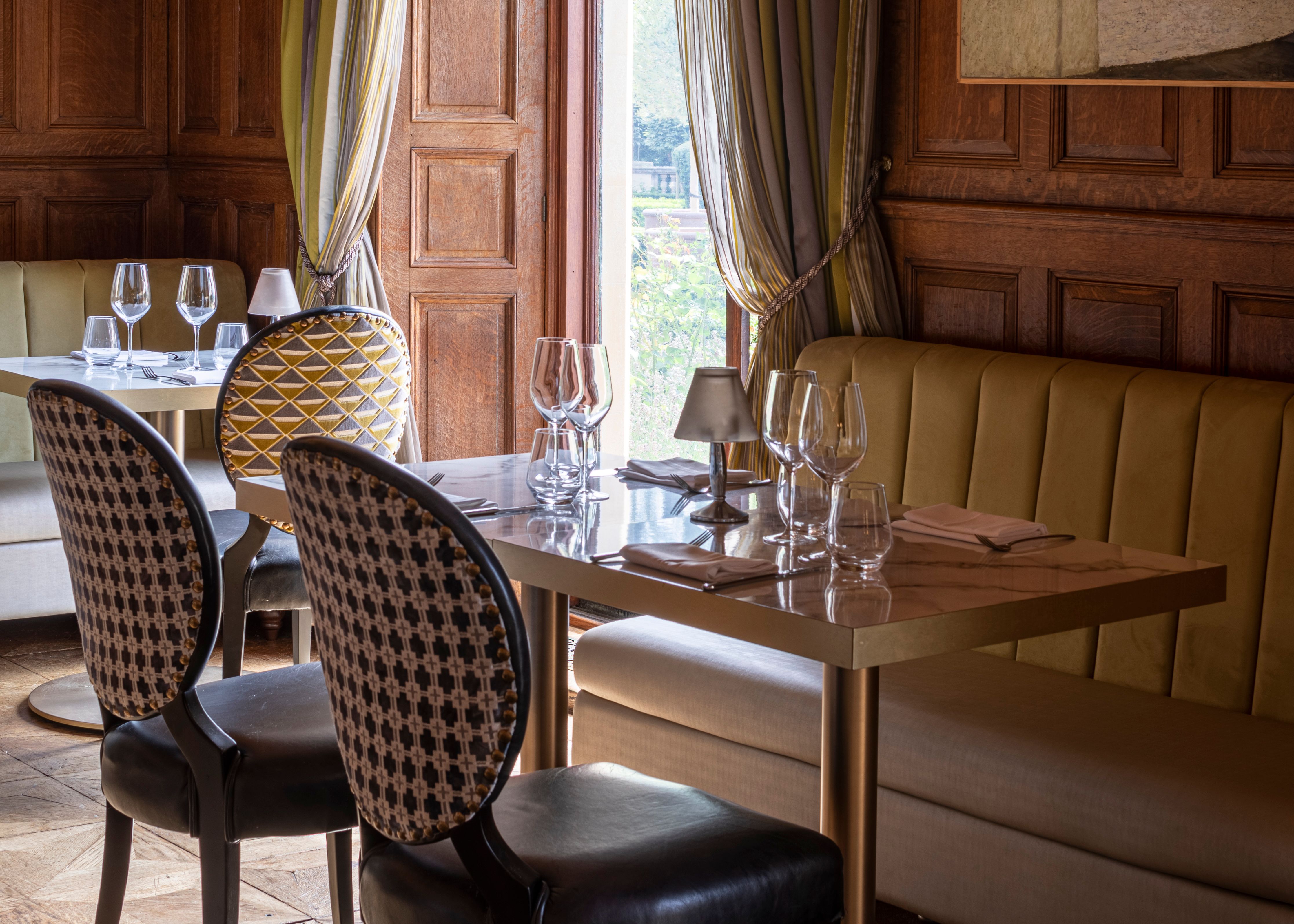 Elegant restaurant interior with wood paneling, upholstered seating, and a set dining table by a window with striped drapes.