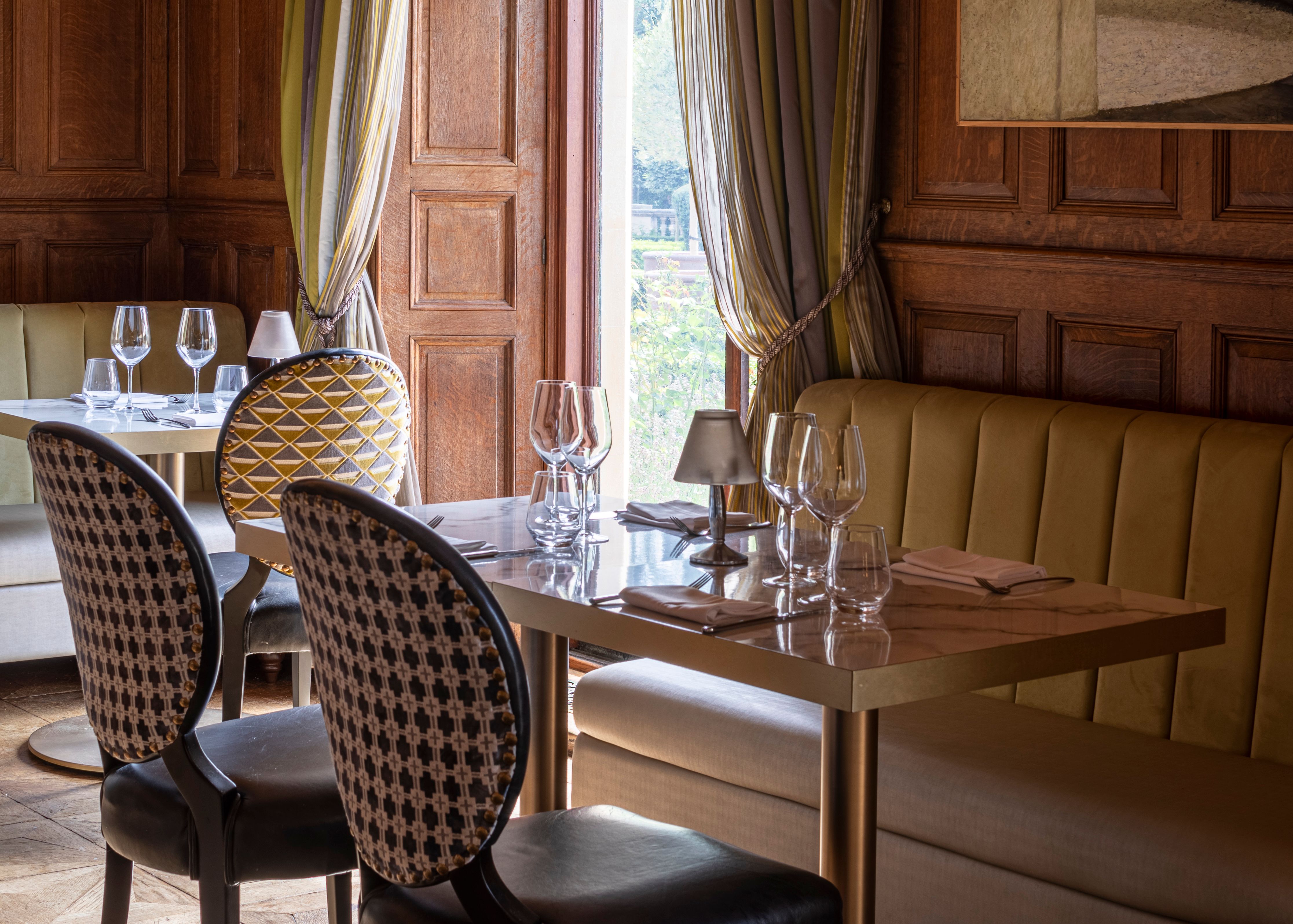 Elegant restaurant interior with wood paneling, upholstered seating, and a set dining table by a window with striped drapes.