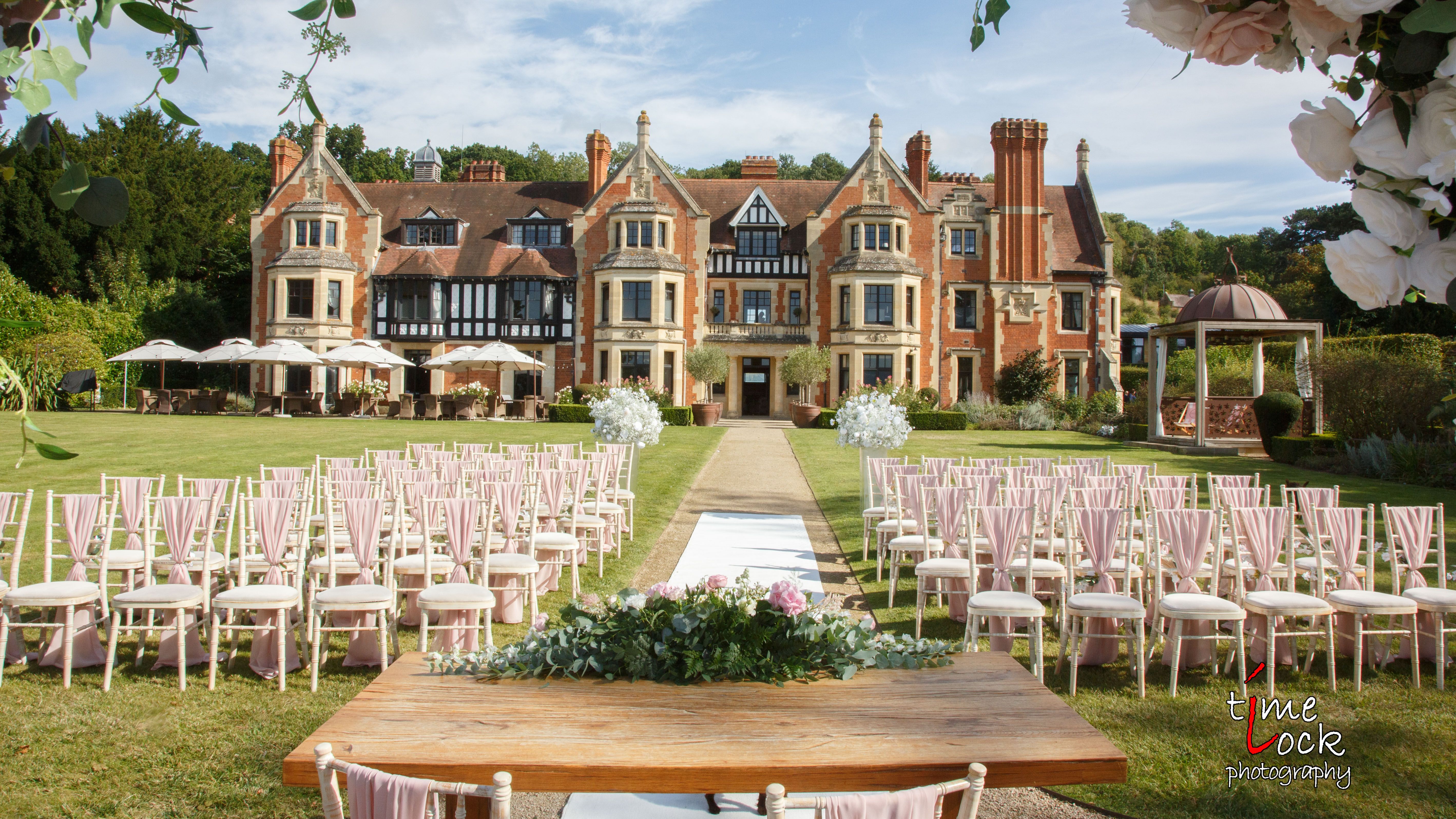 Outdoor wedding ceremony setup with rows of chairs, a decorated table, and a scenic manor house in the background.