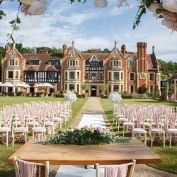 Outdoor wedding ceremony setup with rows of chairs, a decorated table, and a scenic manor house in the background.