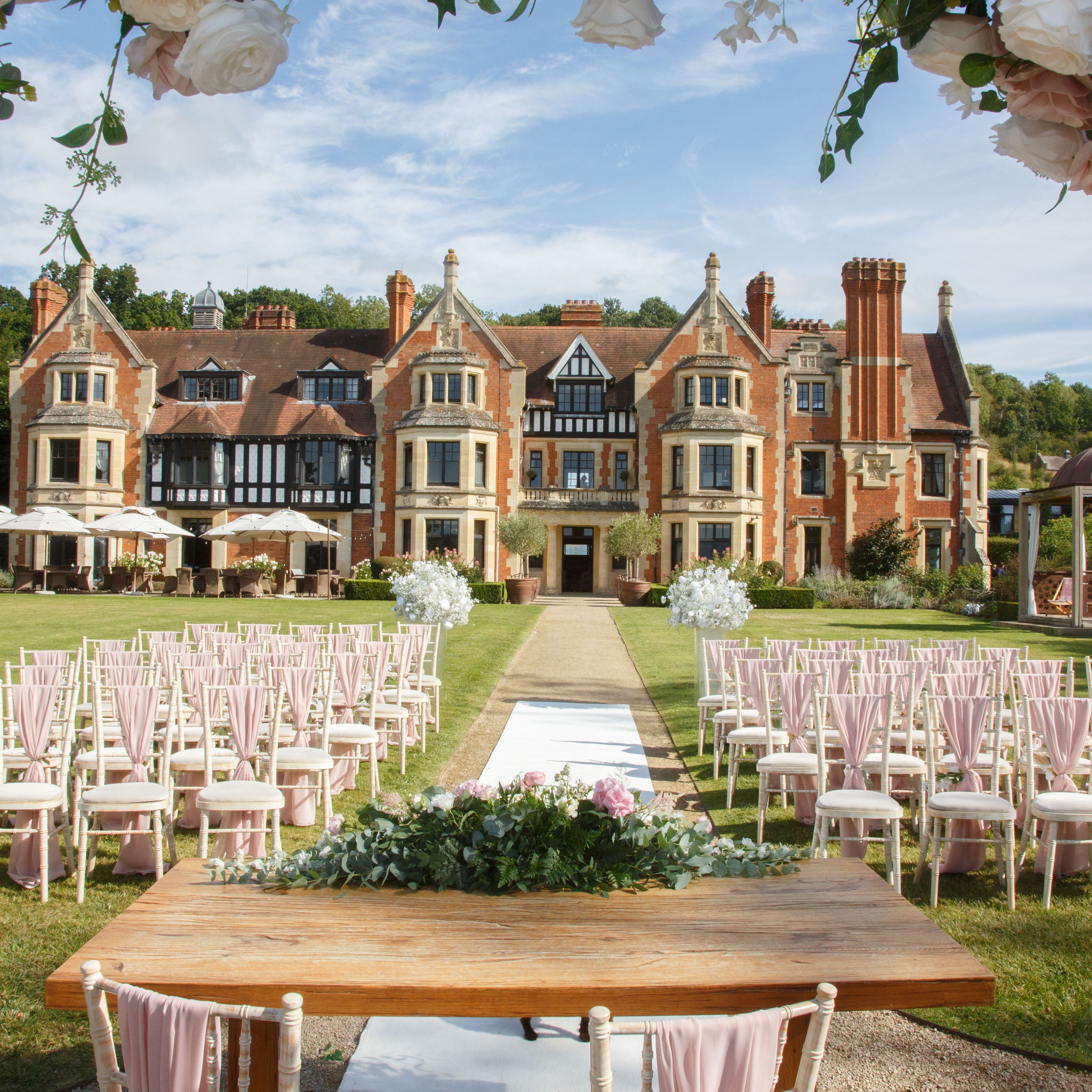Outdoor wedding ceremony setup with rows of chairs, a decorated table, and a scenic manor house in the background.