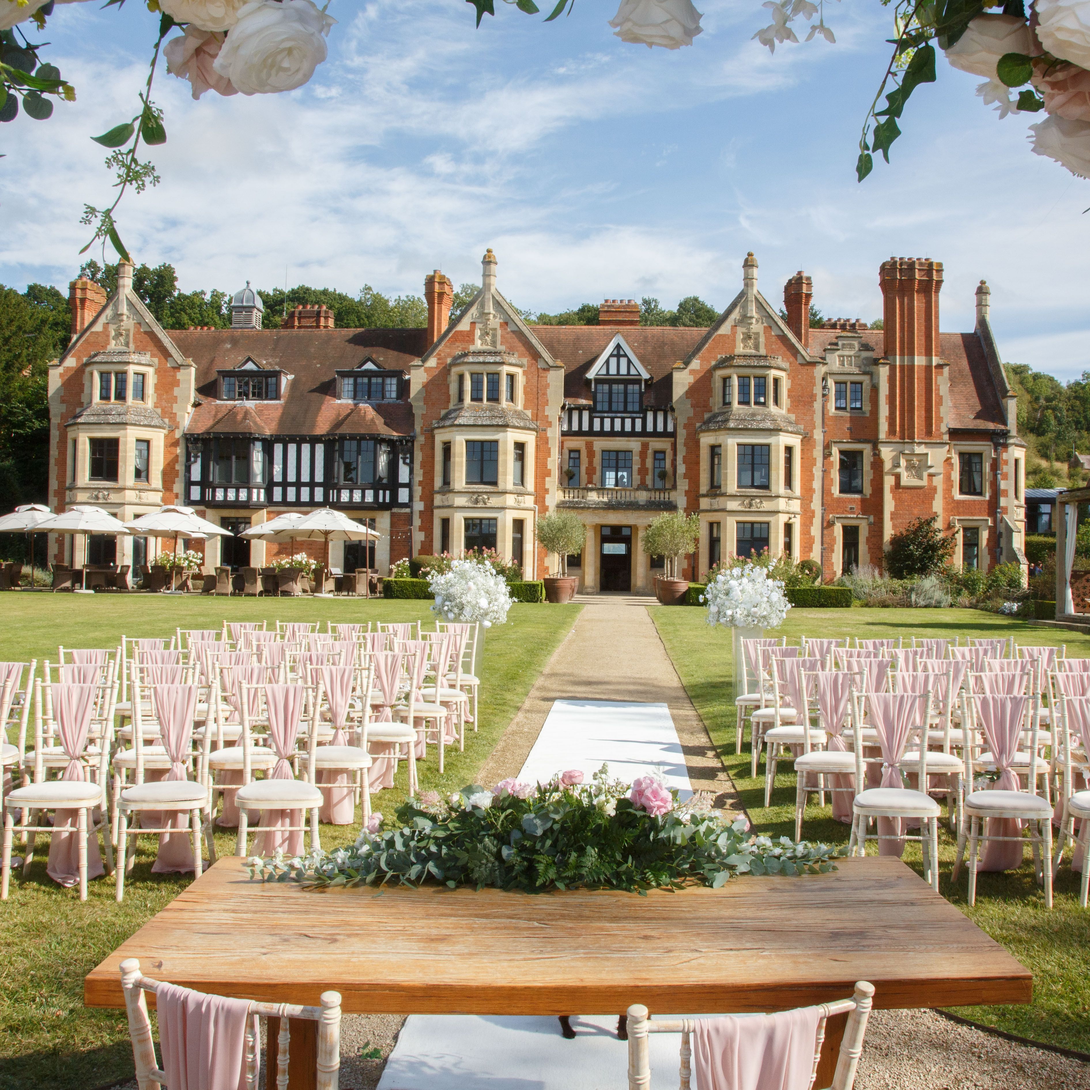 Outdoor wedding ceremony setup with rows of chairs, a decorated table, and a scenic manor house in the background.