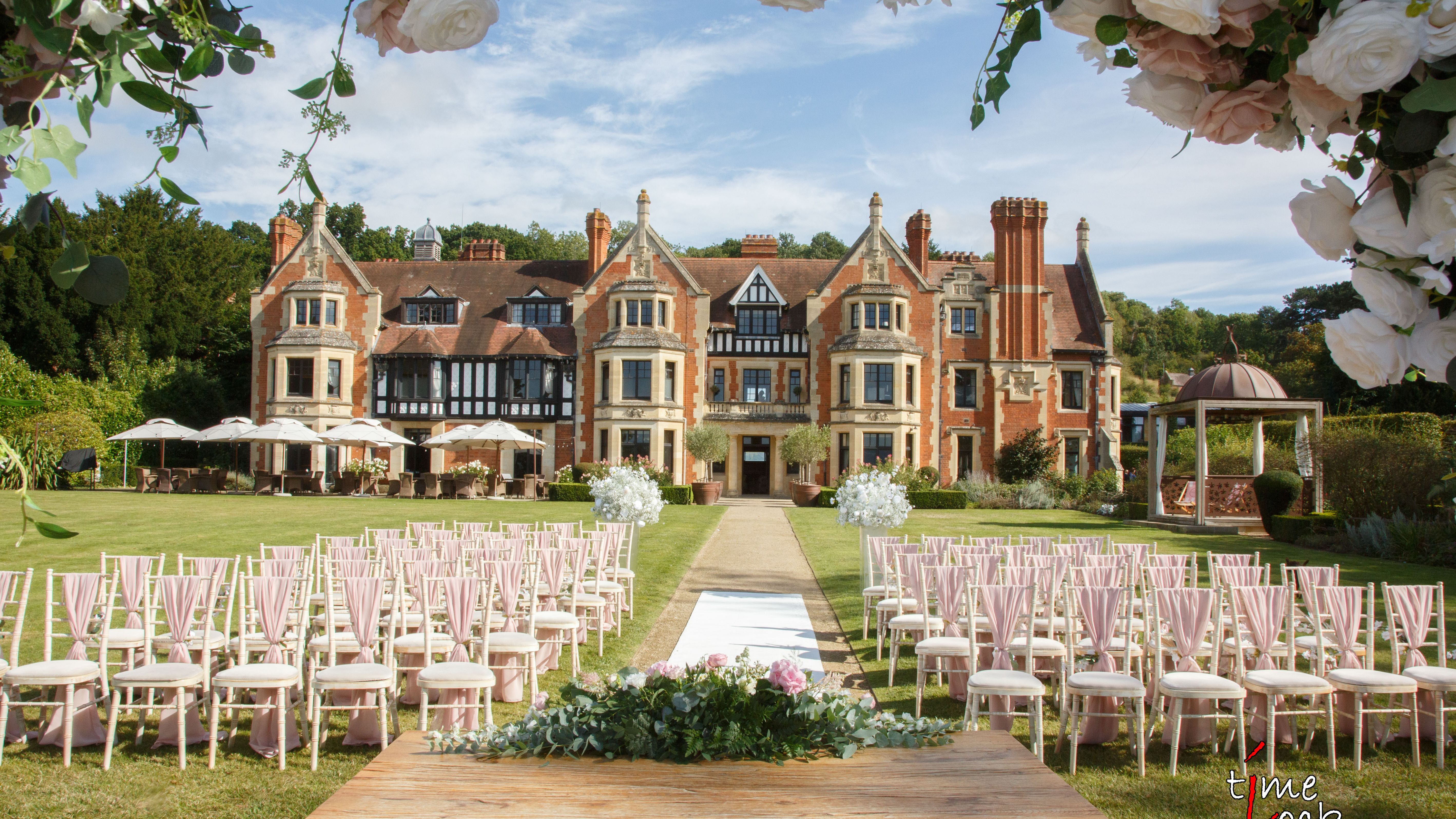 Outdoor wedding ceremony setup with rows of chairs, a decorated table, and a scenic manor house in the background.