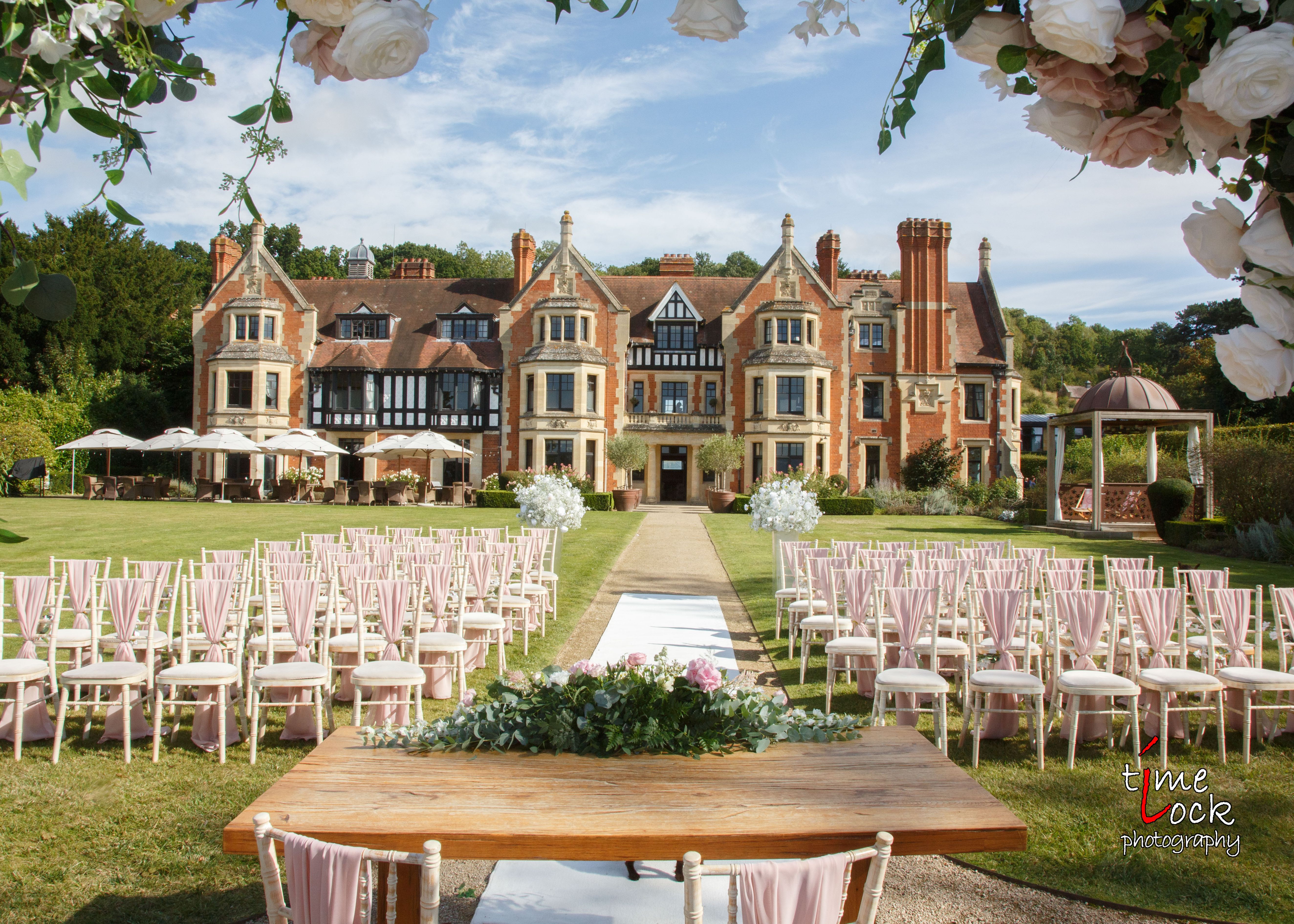 Outdoor wedding ceremony setup with rows of chairs, a decorated table, and a scenic manor house in the background.