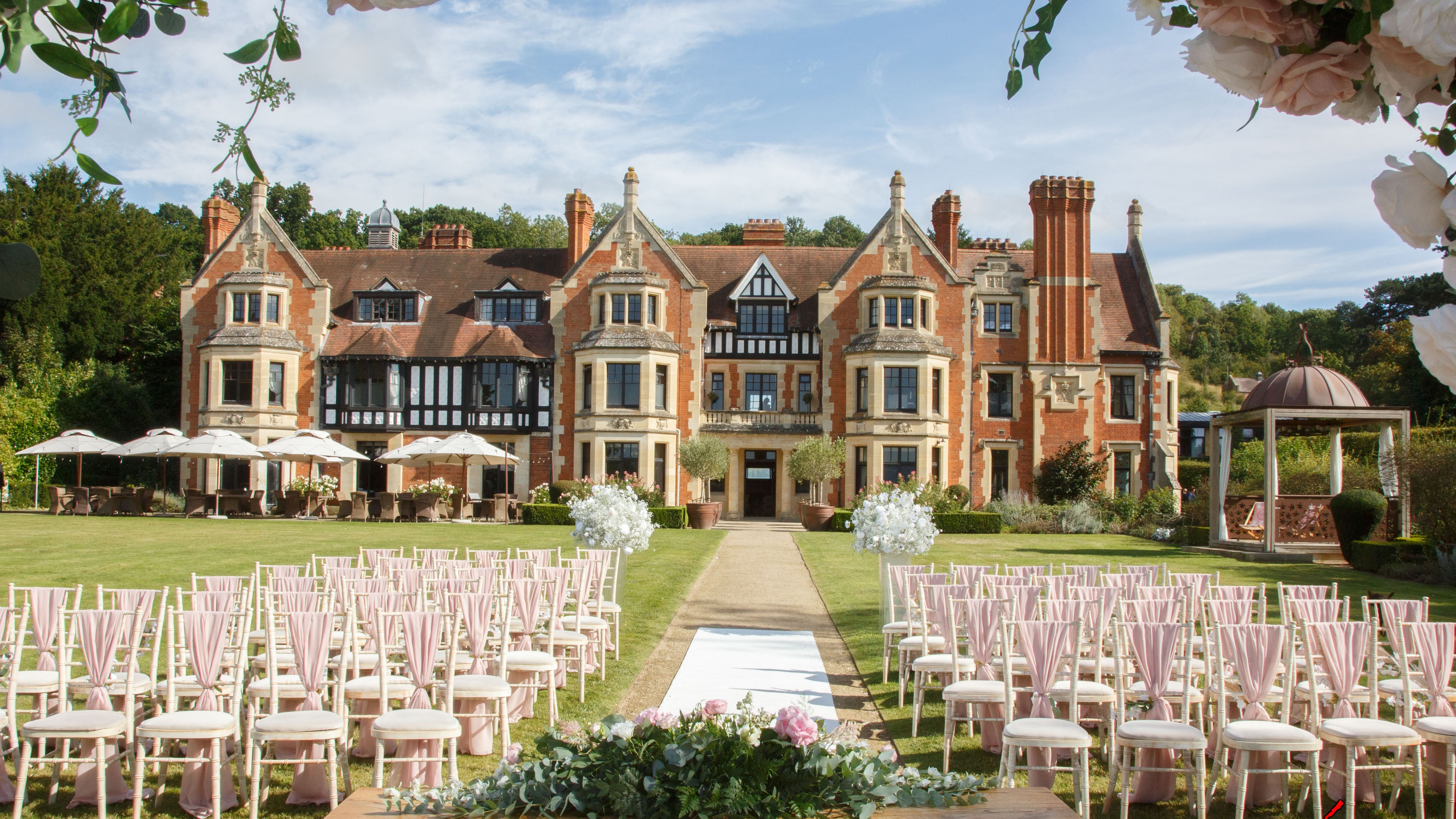 Outdoor wedding ceremony setup with rows of chairs, a decorated table, and a scenic manor house in the background.