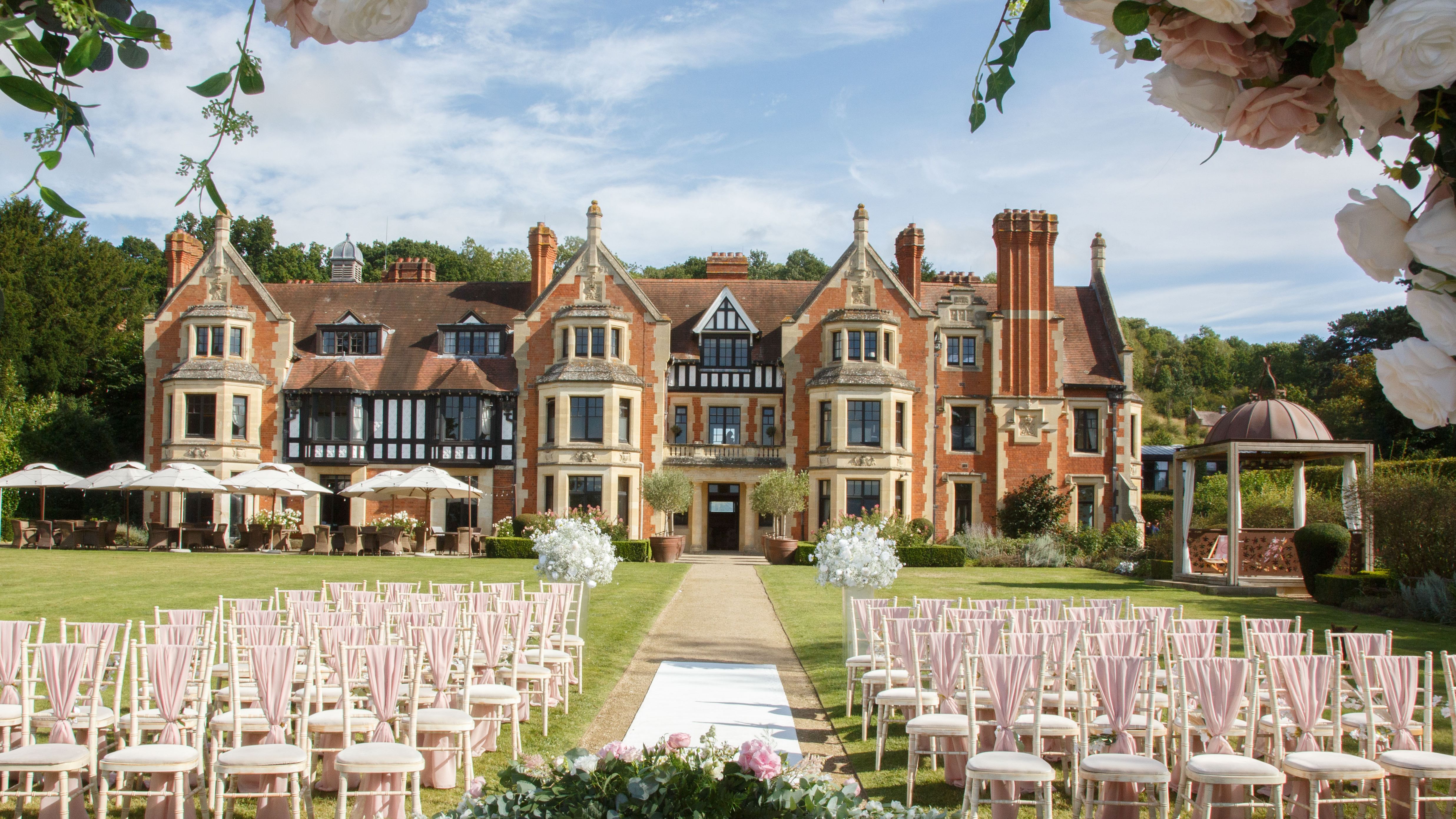 Outdoor wedding ceremony setup with rows of chairs, a decorated table, and a scenic manor house in the background.
