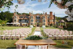 Outdoor wedding ceremony setup with rows of chairs, a decorated table, and a scenic manor house in the background.