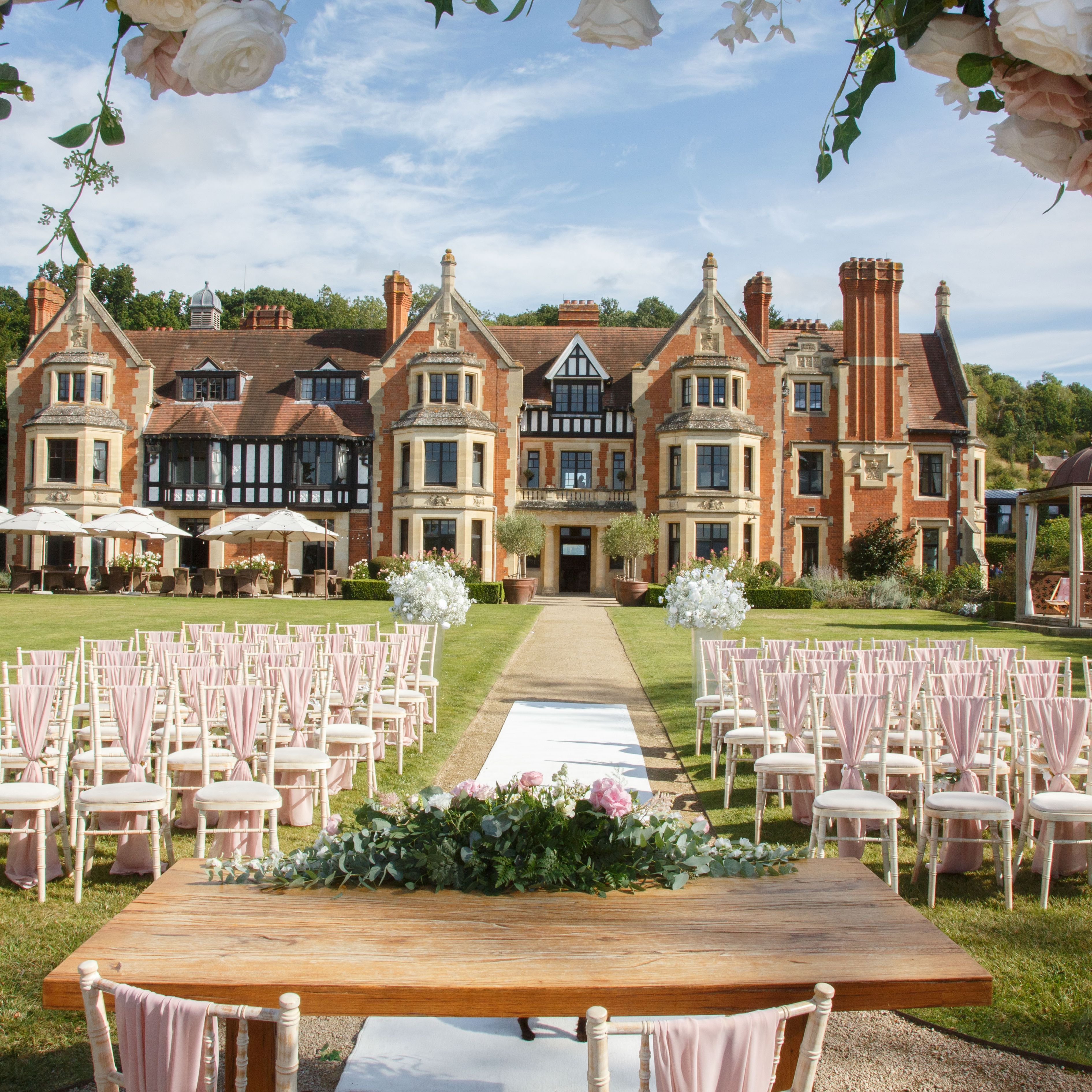 Outdoor wedding ceremony setup with rows of chairs, a decorated table, and a scenic manor house in the background.