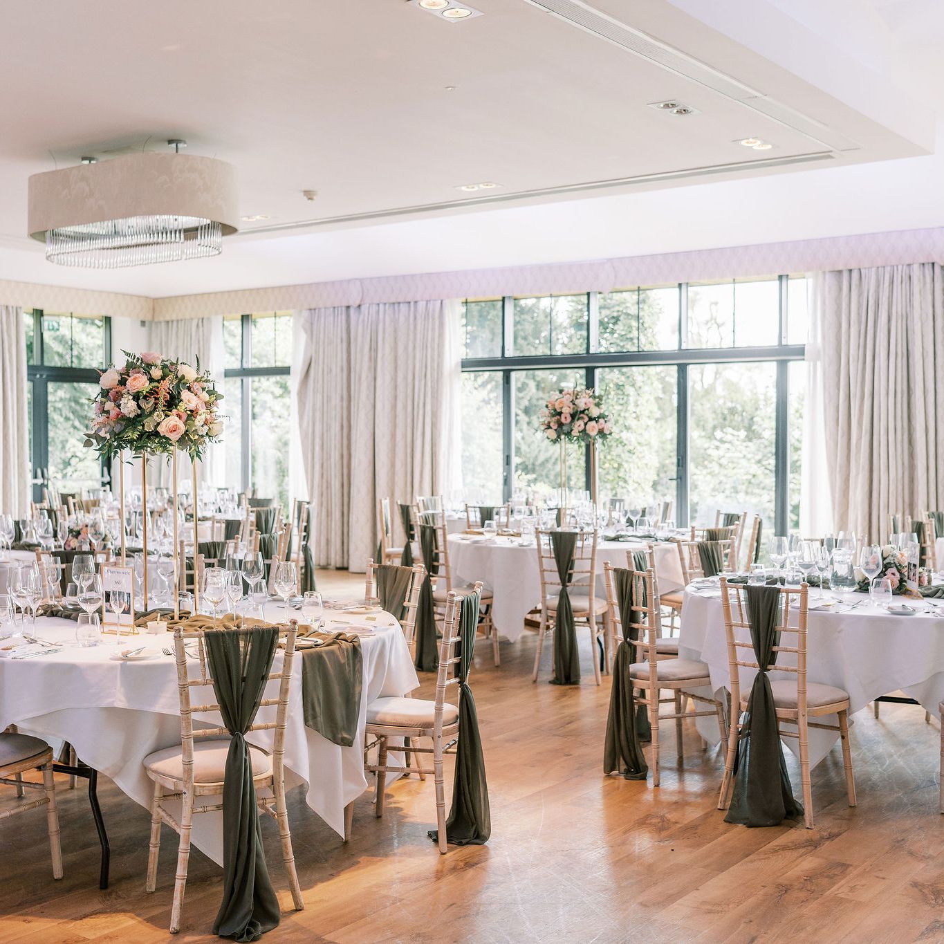 Elegant wedding reception setup with round tables, floral centerpieces, and green chair sashes in a bright venue with large windows.