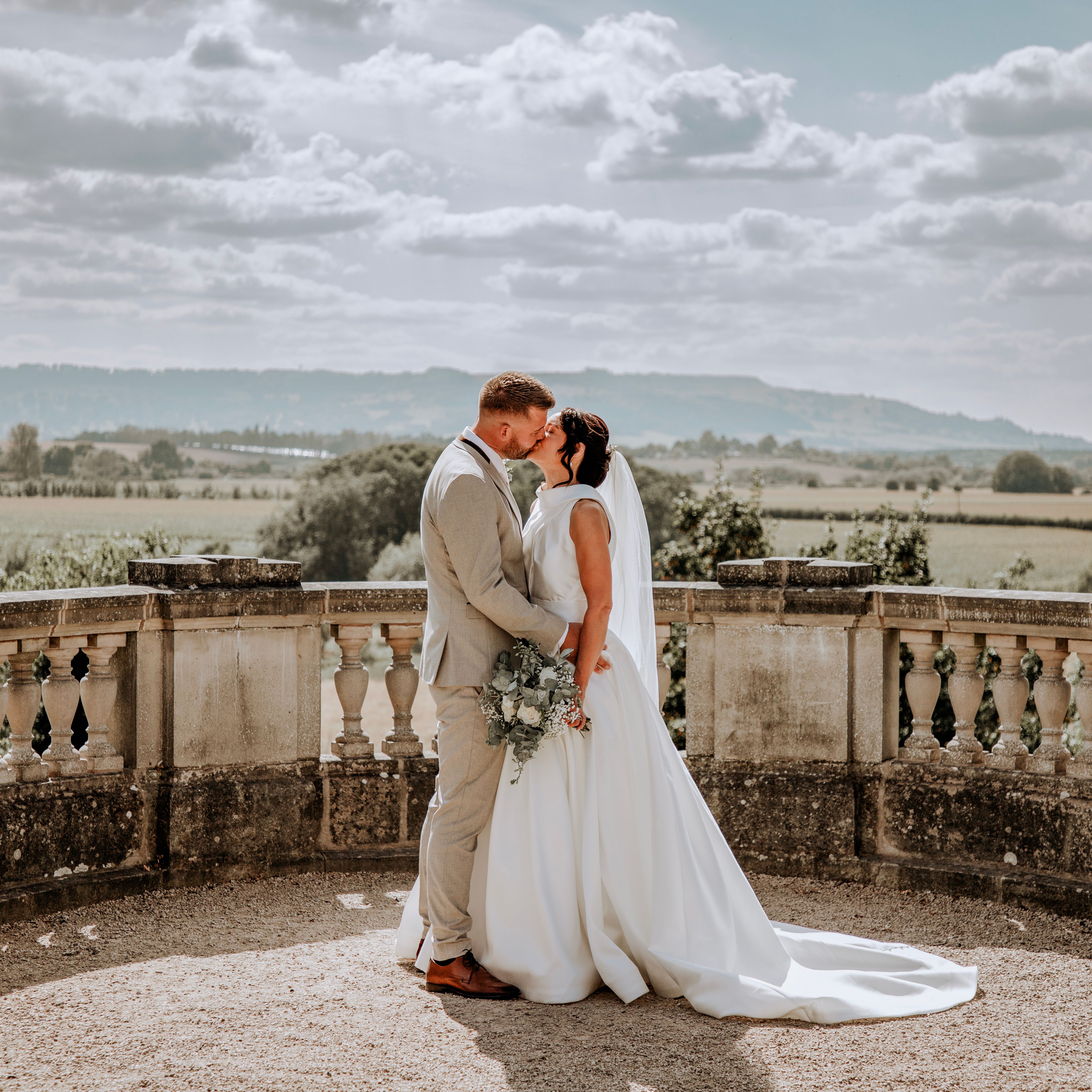 Bride and groom sharing a kiss outdoors on a stone terrace with a scenic countryside background.