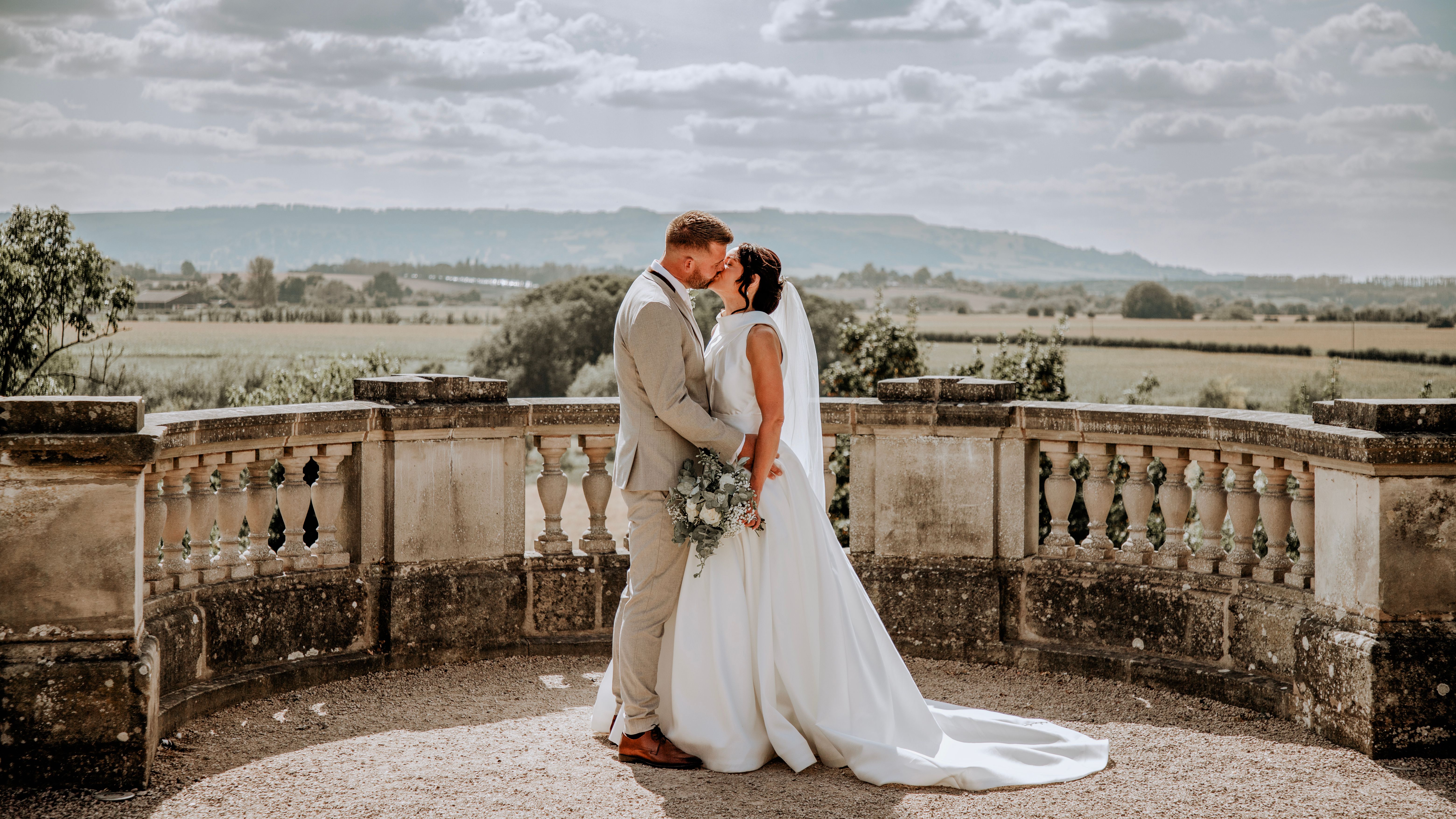Bride and groom sharing a kiss outdoors on a stone terrace with a scenic countryside background.