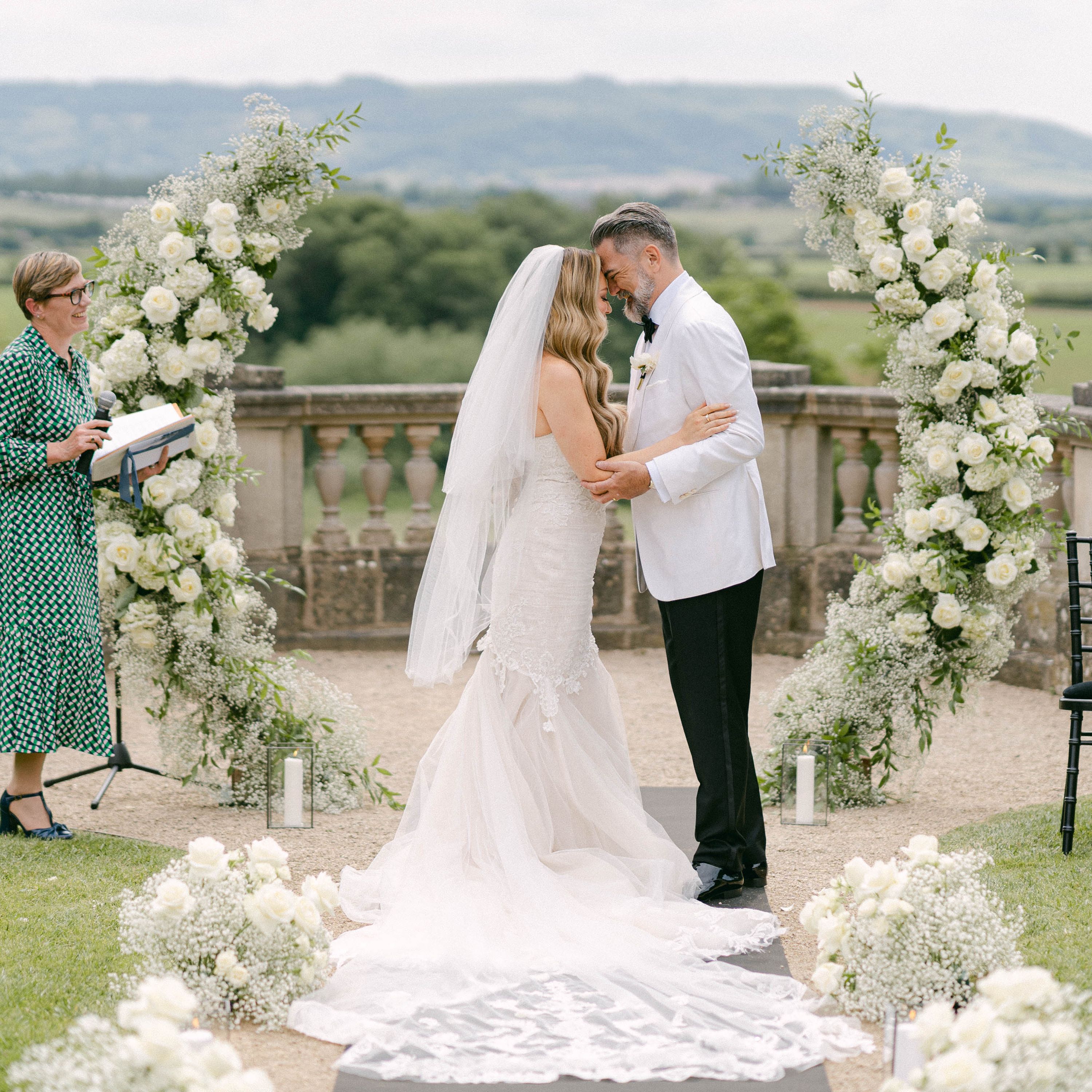 Bride and groom exchanging vows in an outdoor wedding ceremony with floral arches and a scenic countryside backdrop.